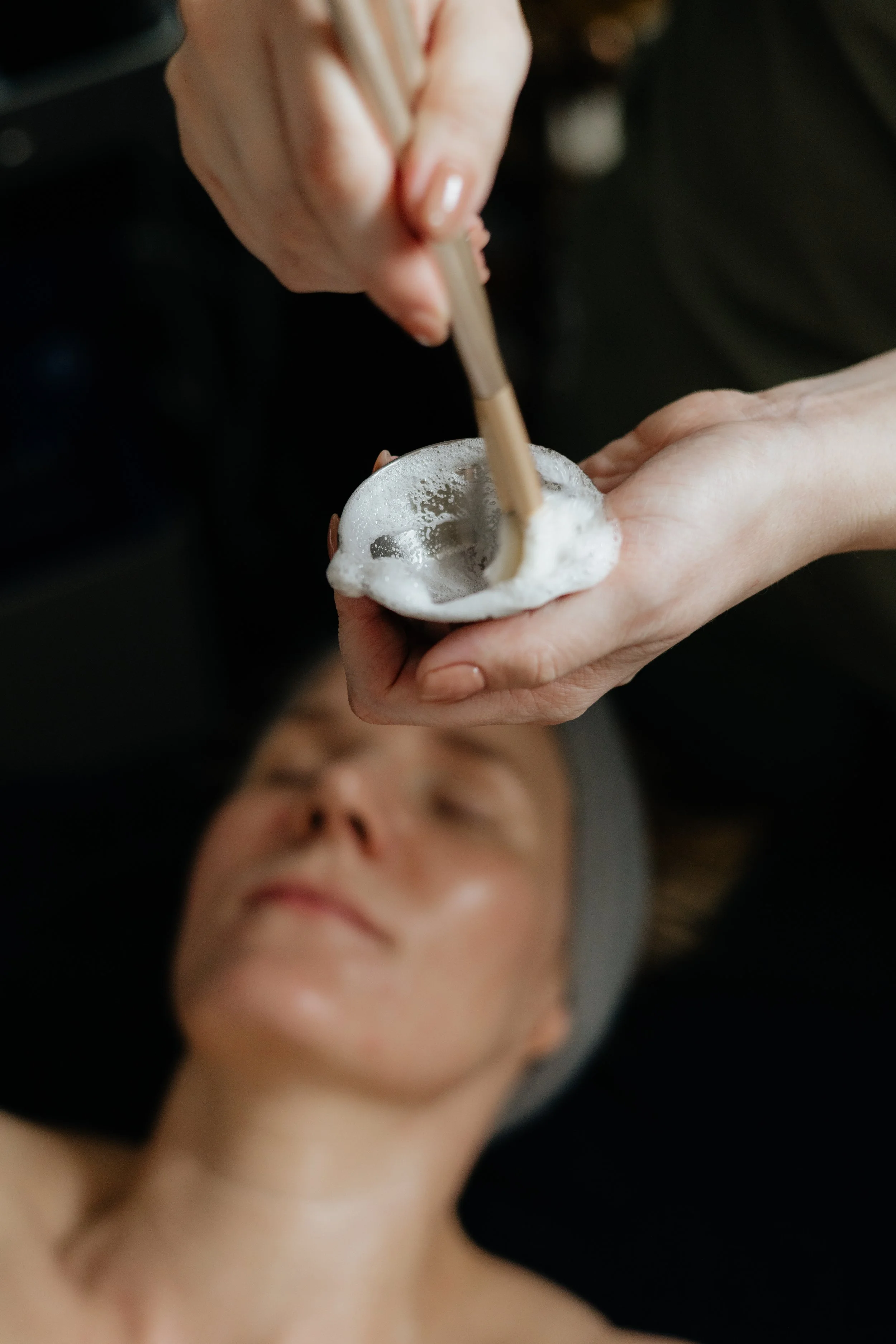 Person receiving a facial treatment, lying down with eyes closed, while another person applies a foam mask or face treatment using a wooden spatula.