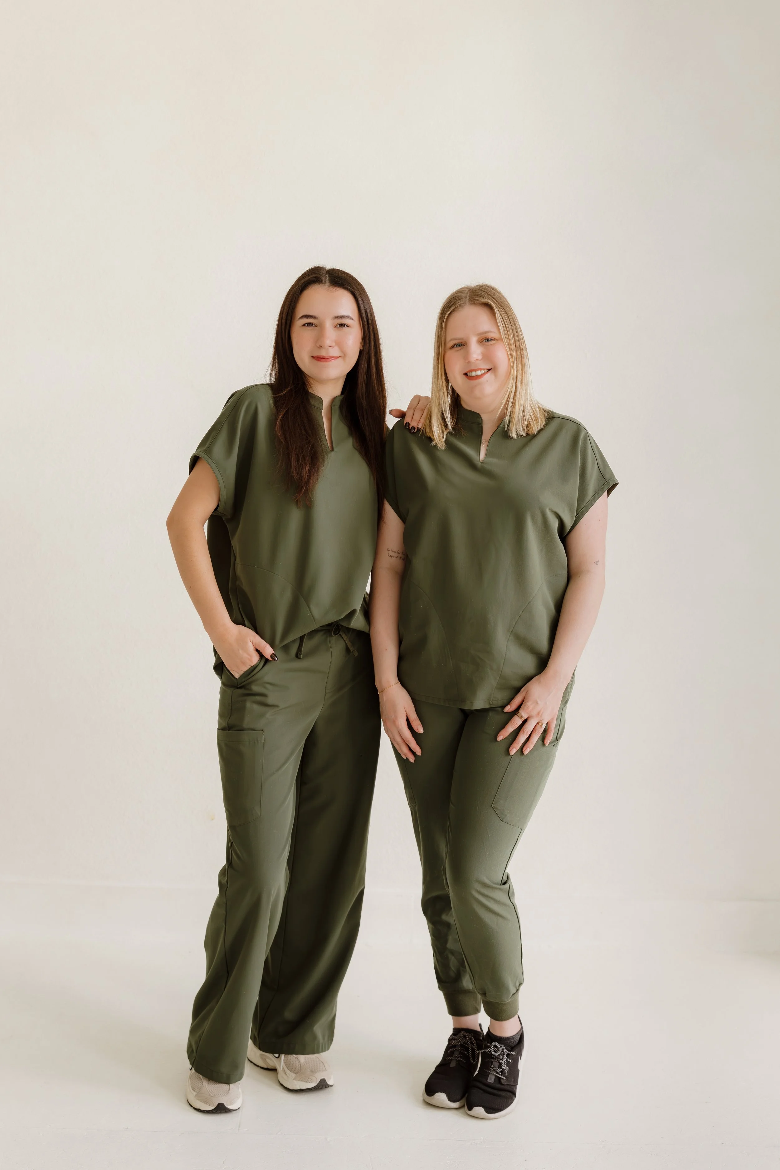 Two women standing side by side dressed in matching khaki-green outfits, against a plain white background. One has long dark hair and the other has shoulder-length blonde hair, both smiling at the camera.