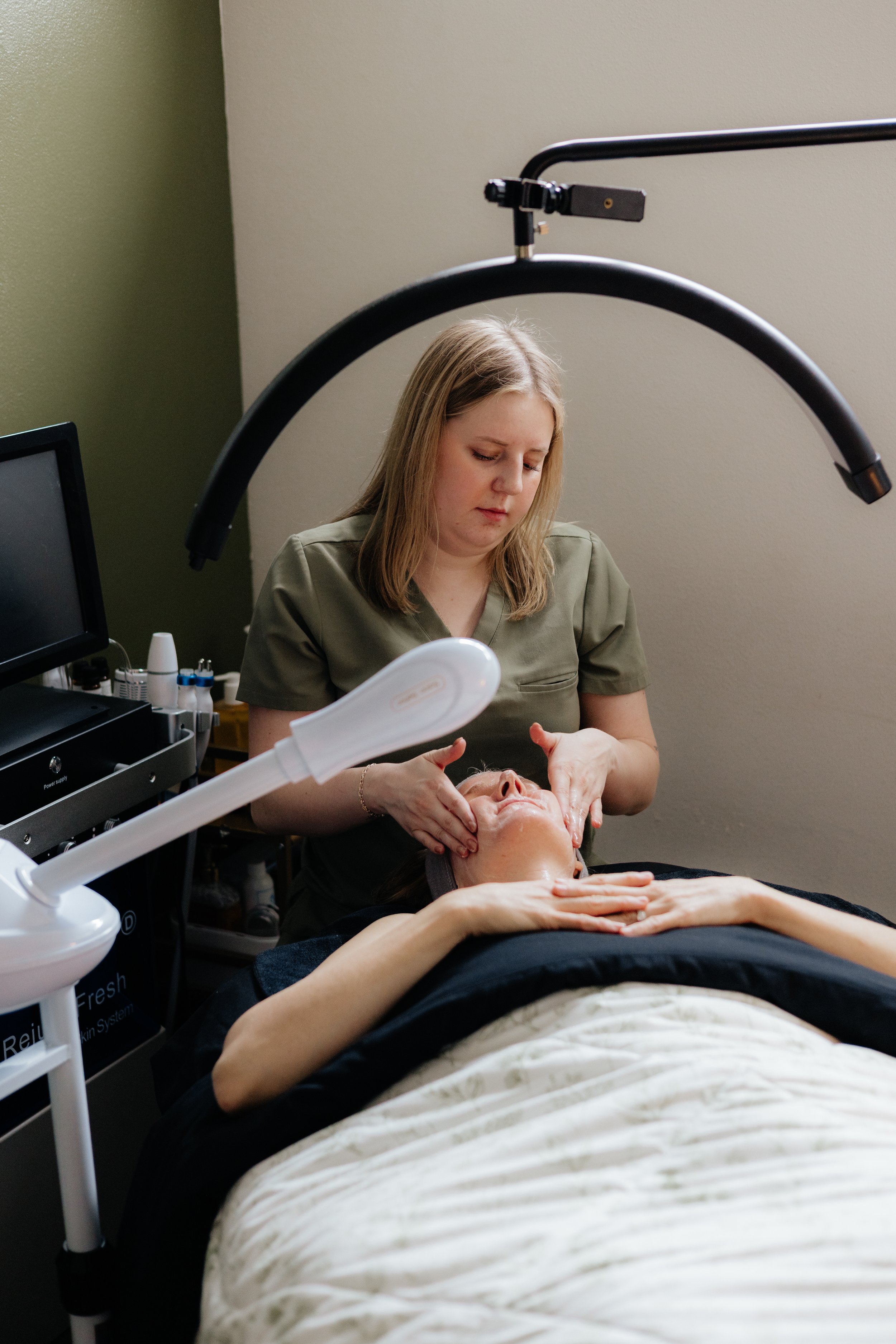 A woman receiving a facial treatment from a skincare professional in a clinic or spa room. The professional is applying a mask or treatment onto the woman's face while she lies on a treatment bed, with various skincare products and equipment nearby.