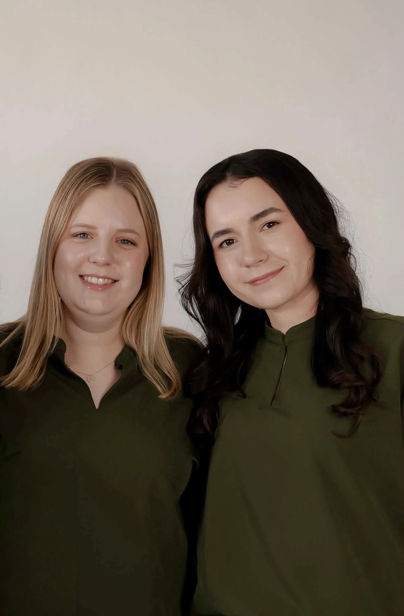 Two women with long hair smiling and standing close, wearing dark green tops against a light background.