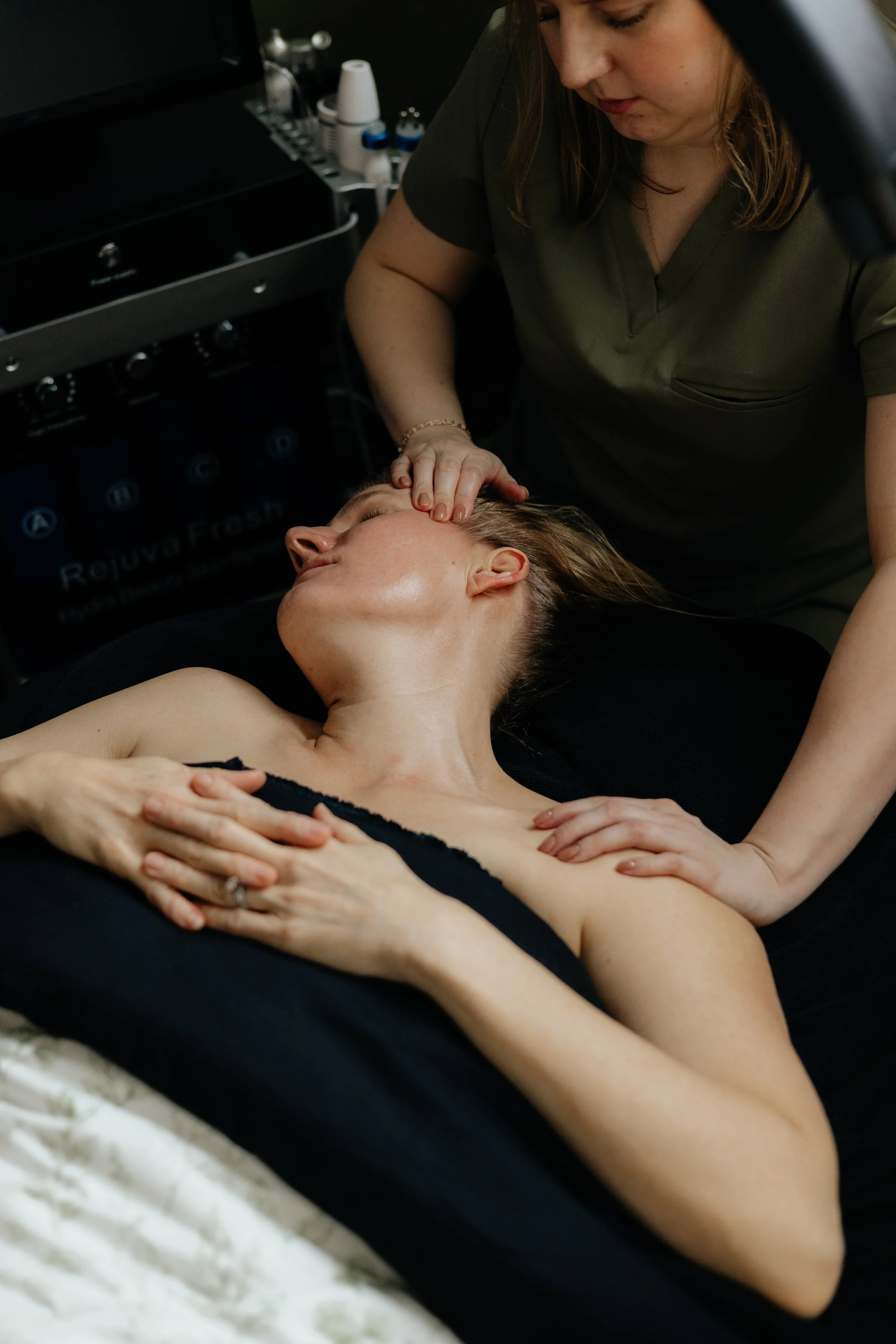 A woman receiving a facial massage from a skincare professional in a spa or clinic setting, with skincare products in the background.