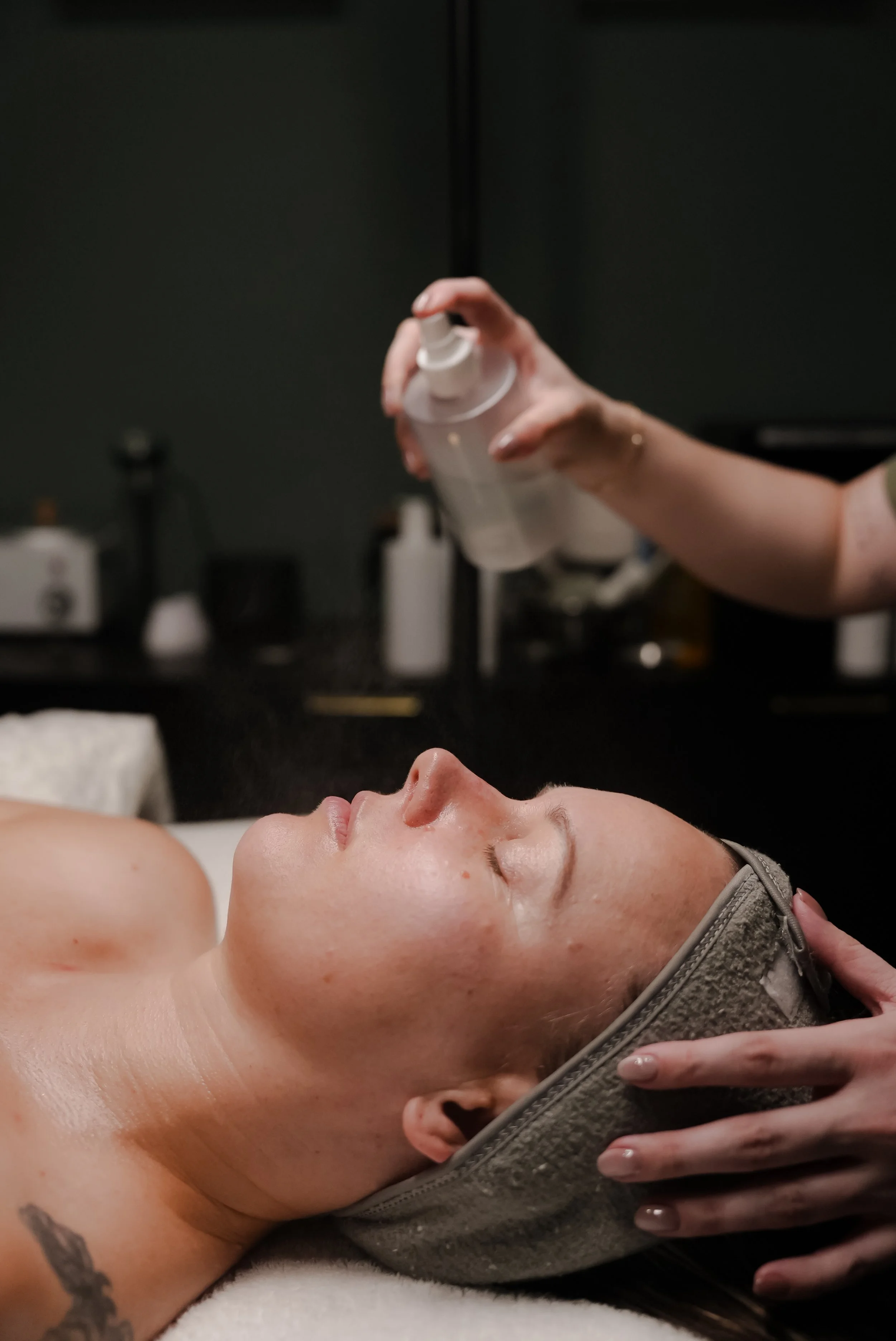A woman lies back on a massage table with eyes closed, receiving facial massage, in a spa or massage therapy room.