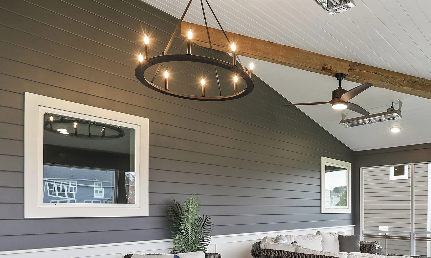 Outdoor porch with ceiling fan, chandelier, gray horizontal siding walls, two small windows with white trim, a white cushioned sofa, and potted plant.