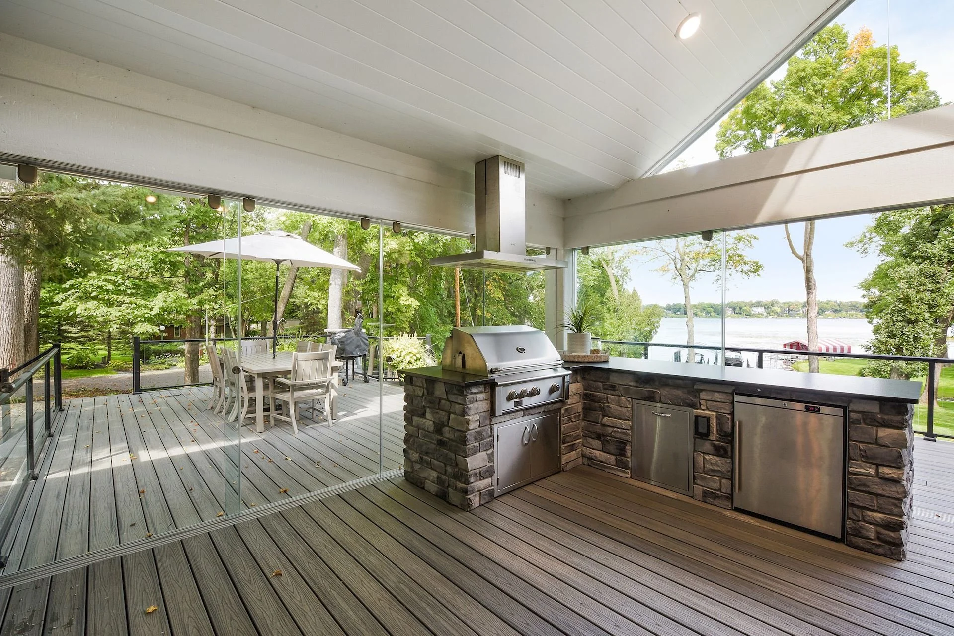 Outdoor screened-in porch with a kitchen area featuring a grill, stone counter, and refrigerator, overlooking a deck with outdoor dining furniture and a lake view surrounded by trees.