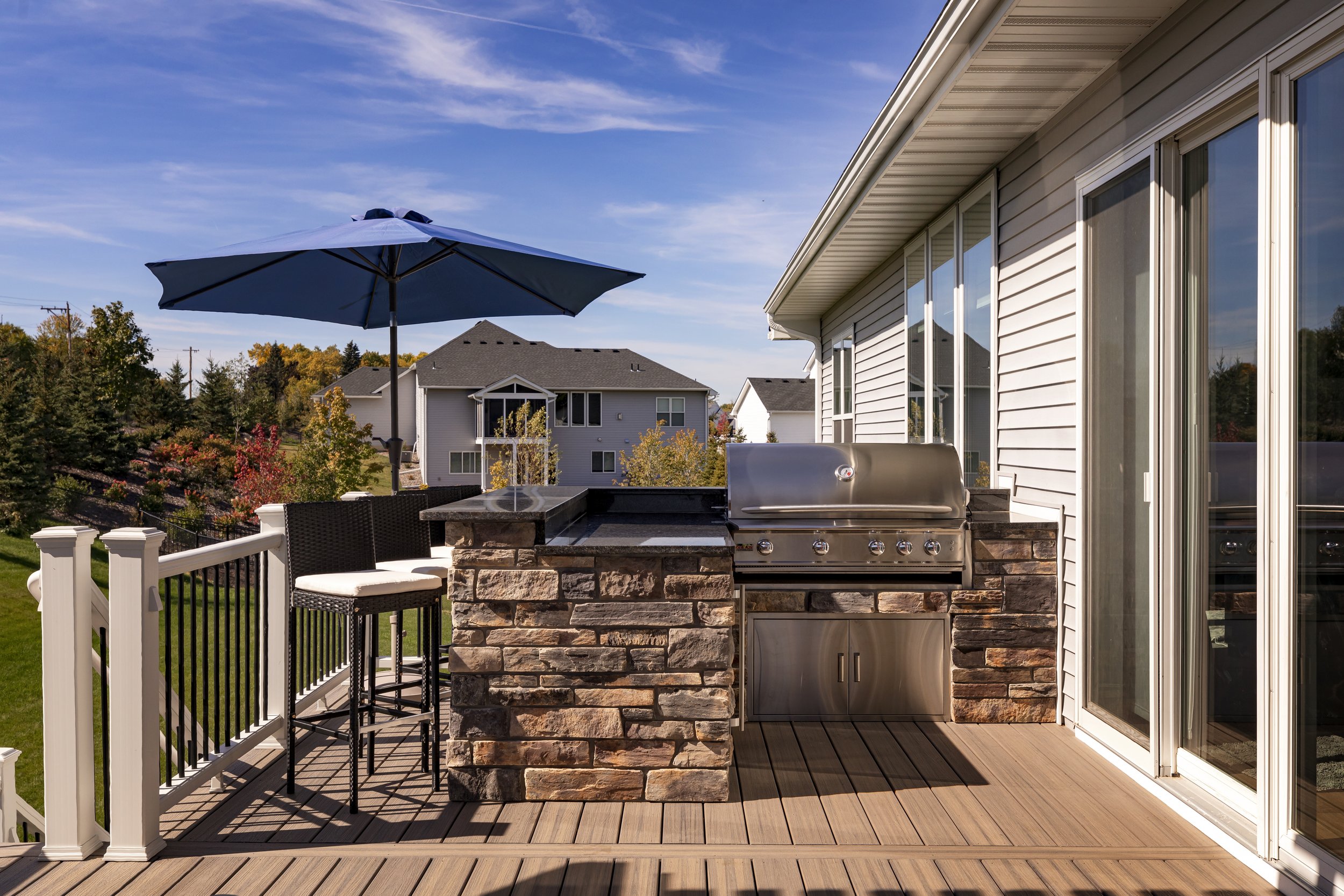 Outdoor balcony with built-in stainless steel gas grill, stone counter and cabinet, black wicker bar stools with white cushions, blue patio umbrella, and view of neighboring houses and trees under a clear blue sky.