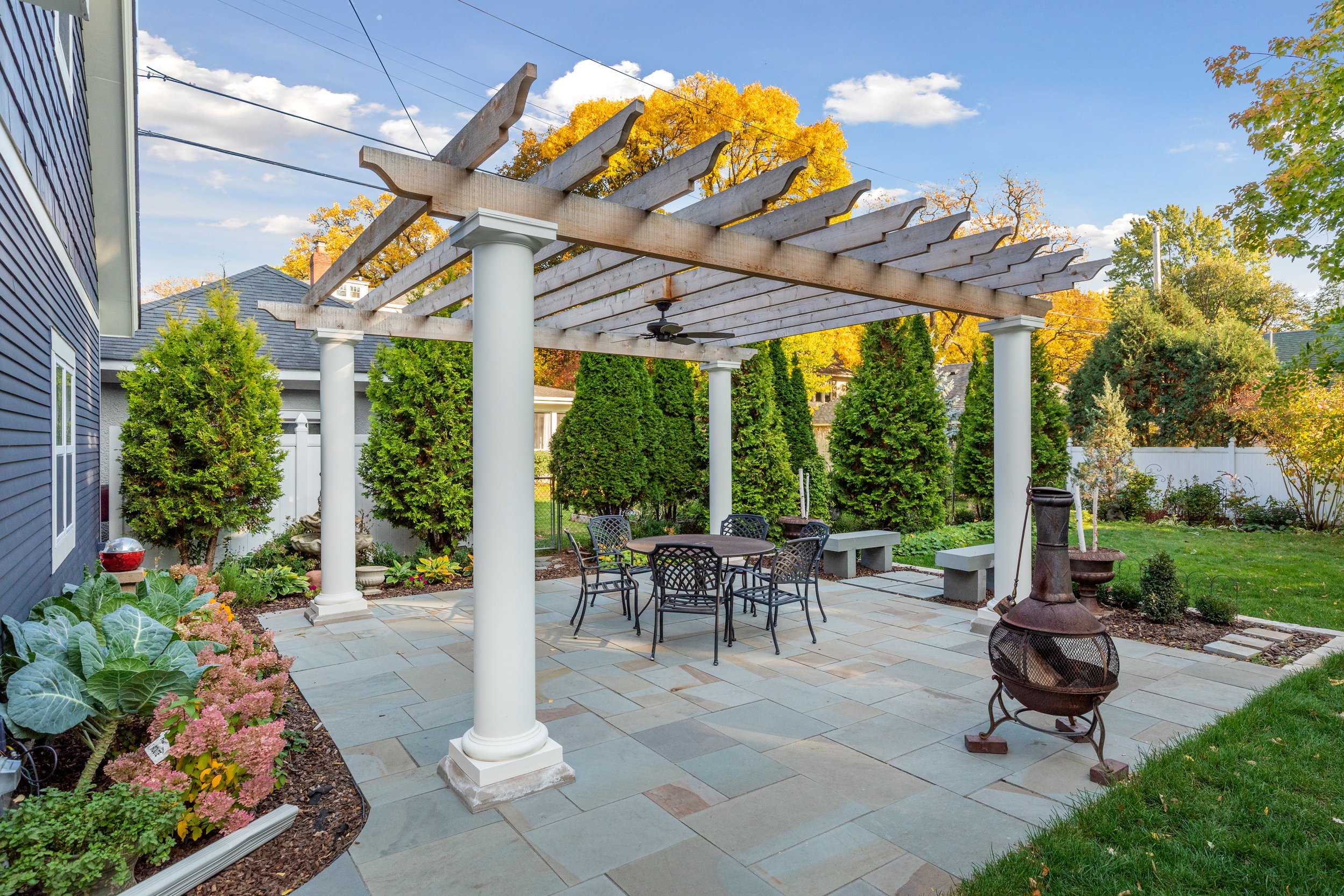 A backyard patio with a pergola, metal outdoor furniture, and a chimney-style fire pit, surrounded by green trees and shrubs under a partly cloudy sky.