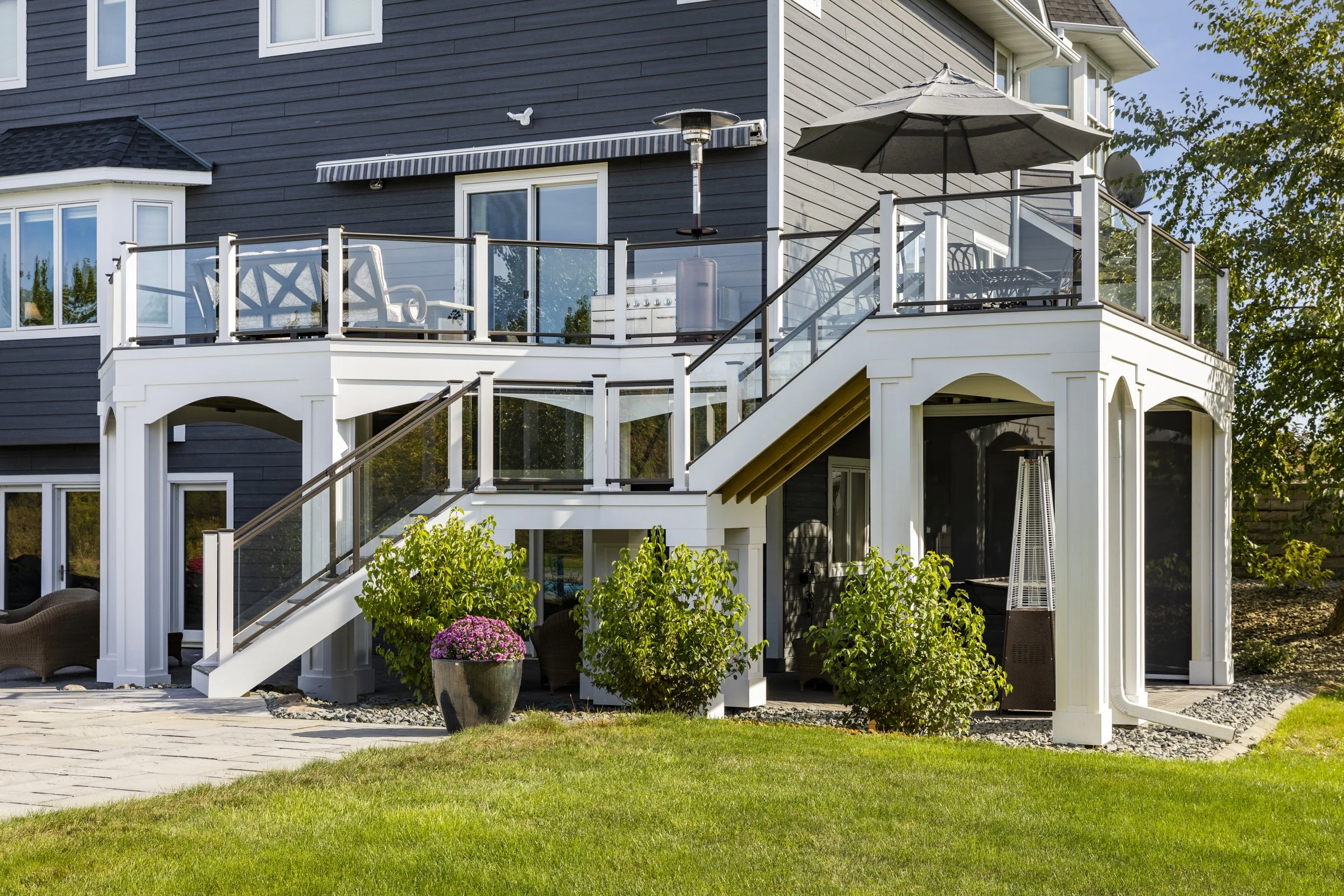 A multi-level deck with glass and white railing, outdoor furniture, an umbrella, a patio heater, potted plants, and a lawn.