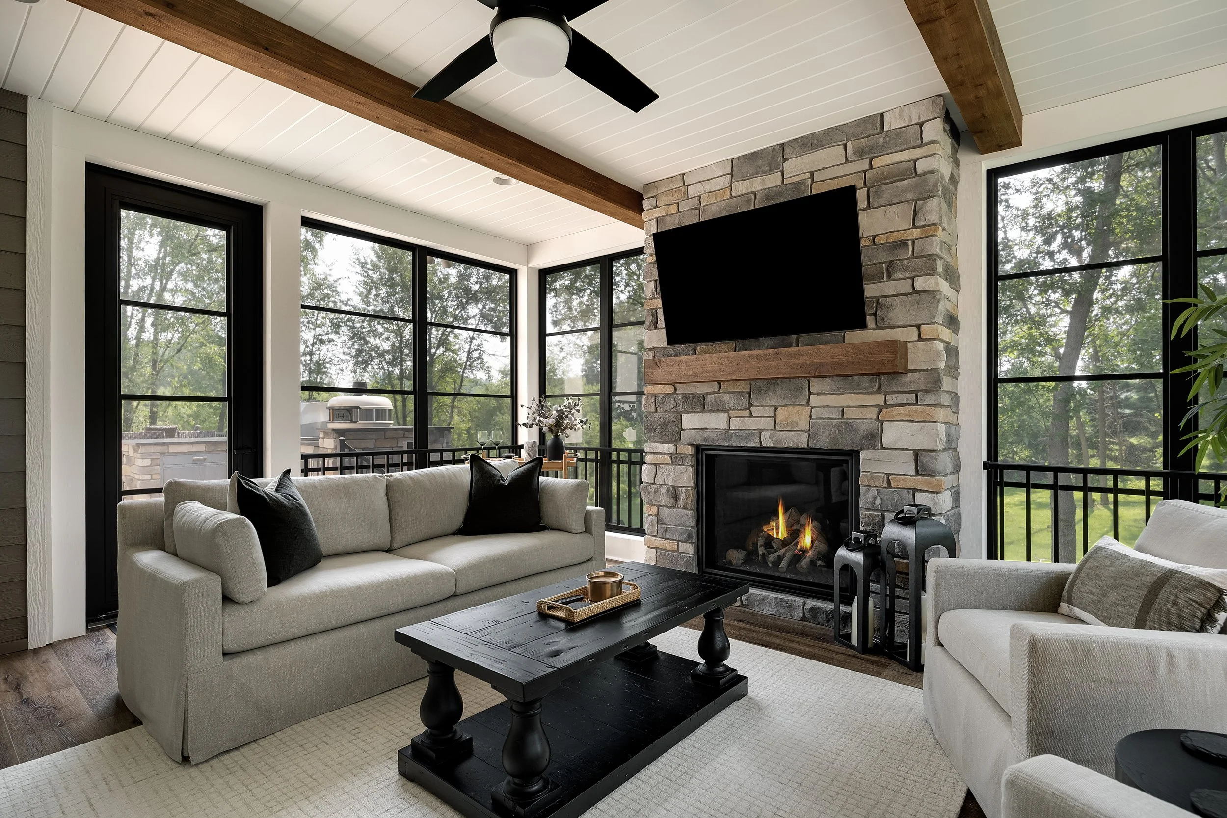 Living room with beige sofas, black coffee table, fireplace, large windows, and mounted TV on a stone wall.