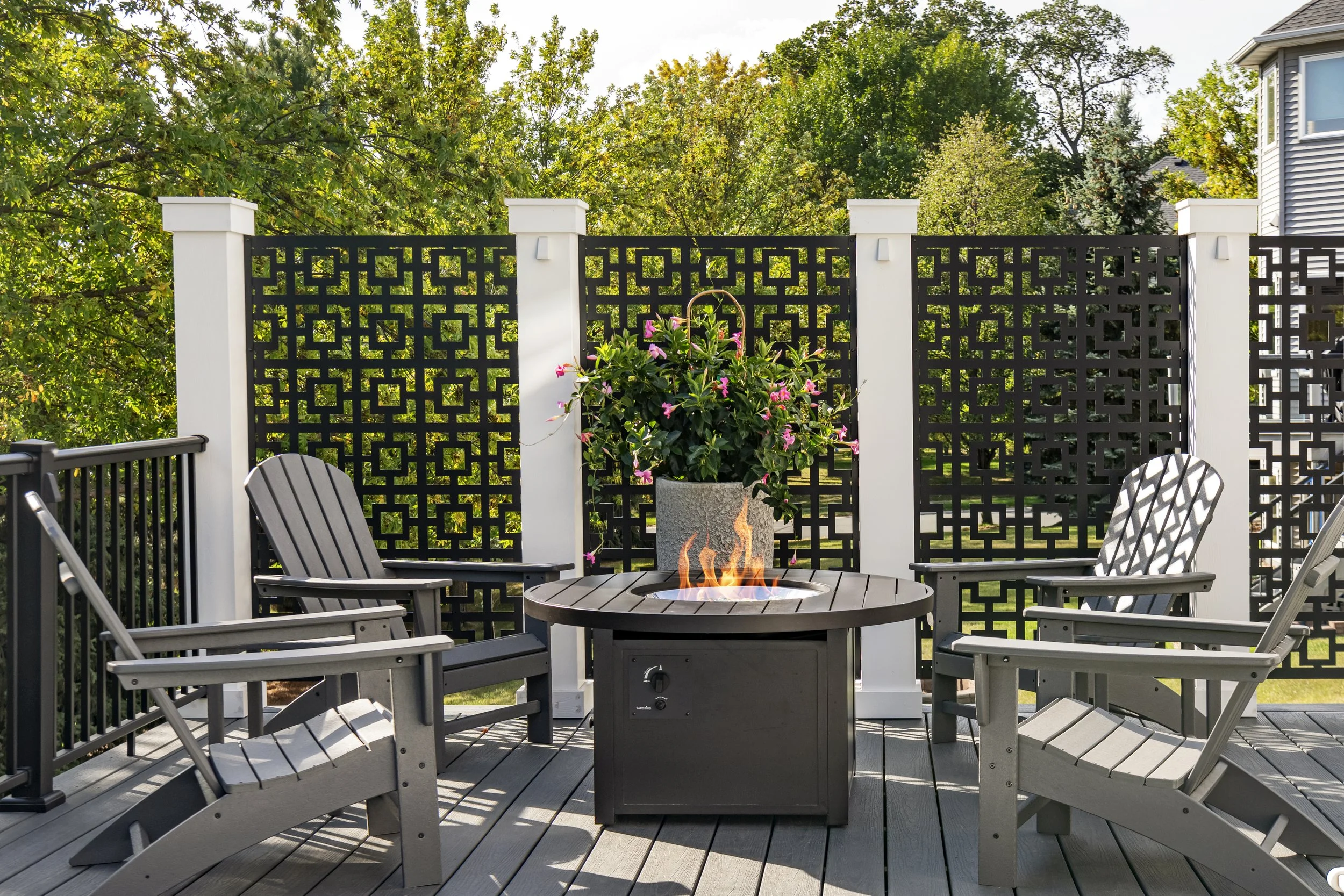 Outdoor patio with four gray Adirondack chairs around a fire pit, decorated with a potted flowering plant, enclosed by a black decorative fence, with lush green trees in the background.