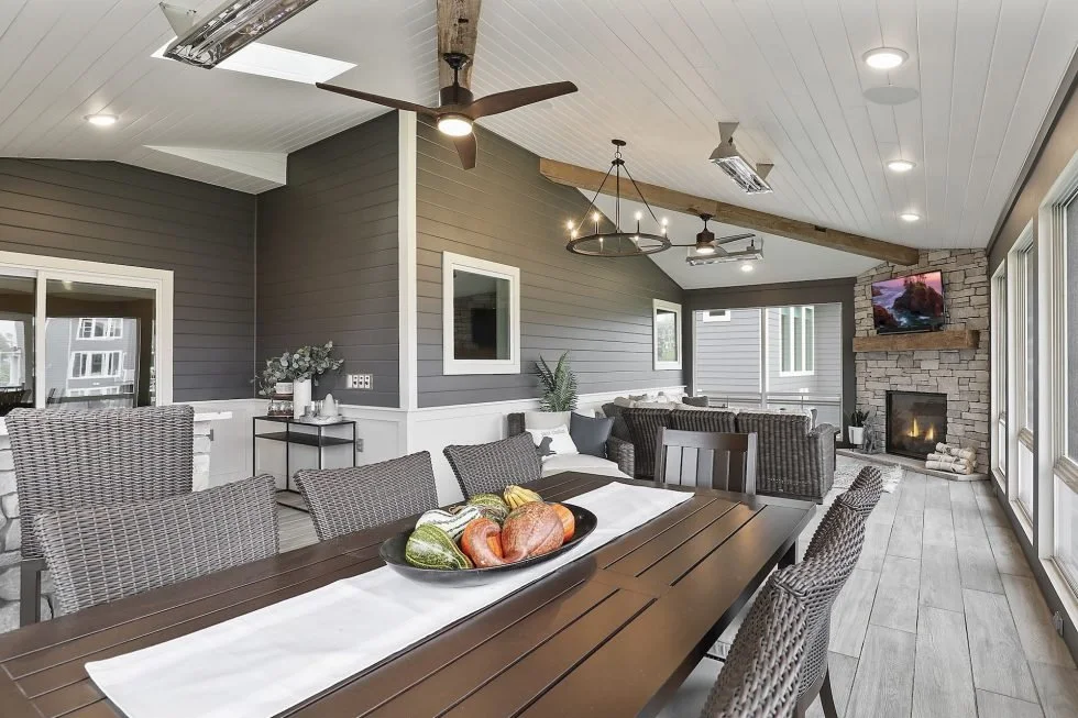 Bright indoor living and dining area with gray walls, white ceiling, ceiling fans, and large windows, featuring a dining table with woven chairs, a cozy seating area with a fireplace, and a mounted television.