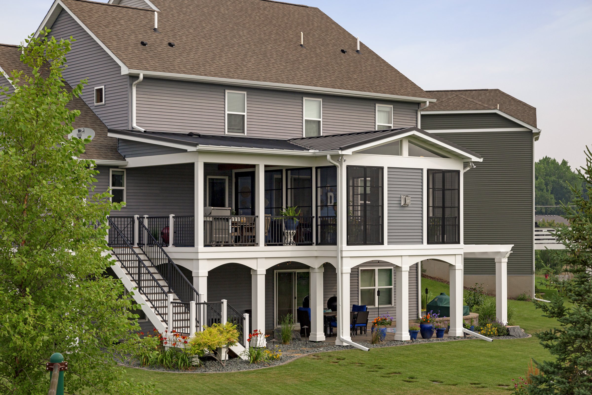 A multi-story house with gray siding, a brown roof, and a large screened-in porch on the upper level, with outdoor furniture and plants.