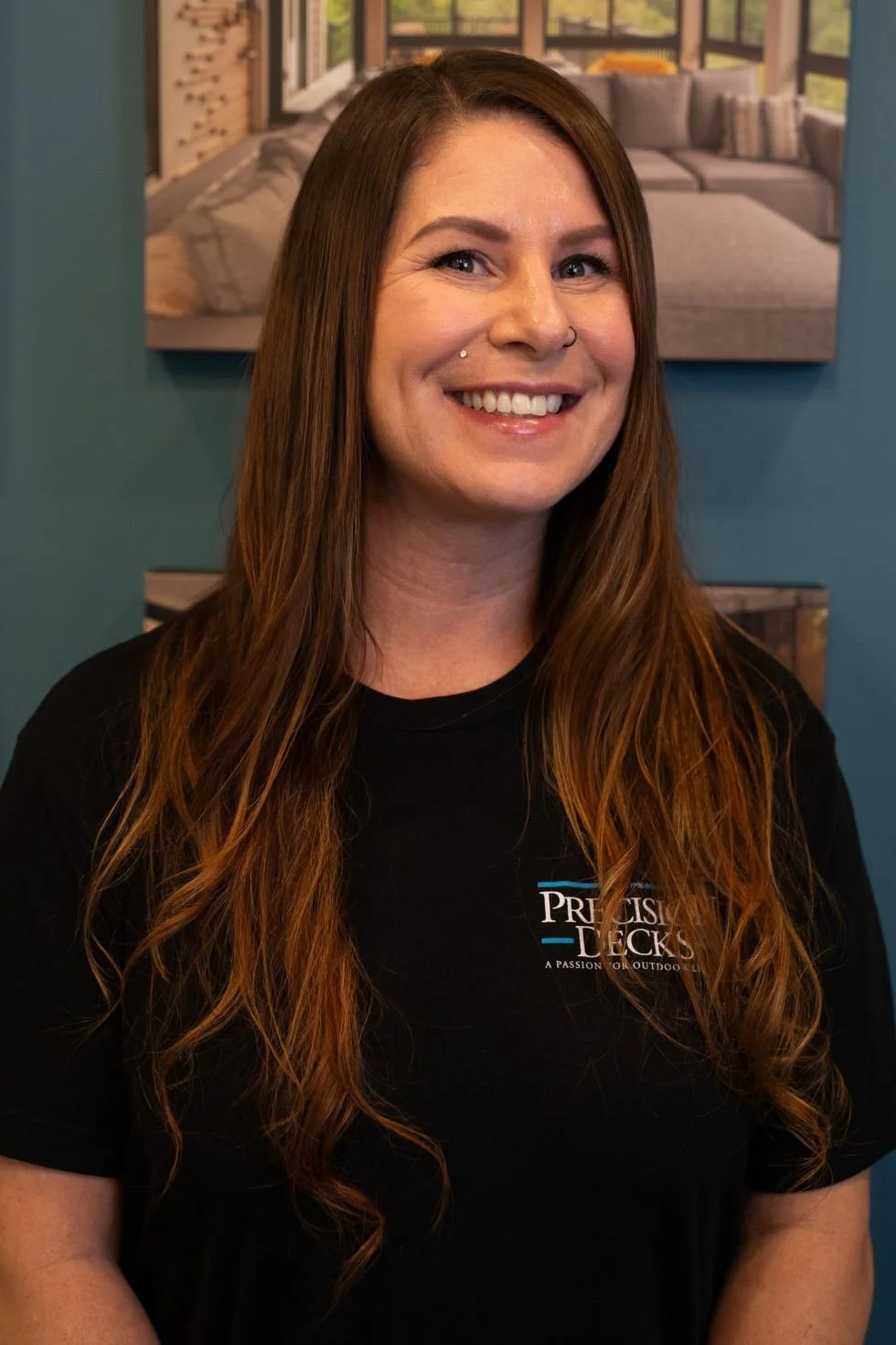 A smiling woman with long red hair and a nose piercing, wearing a black t-shirt with a logo that says 'Precision Decks,' standing in front of a blue wall with artwork of a modern living room.