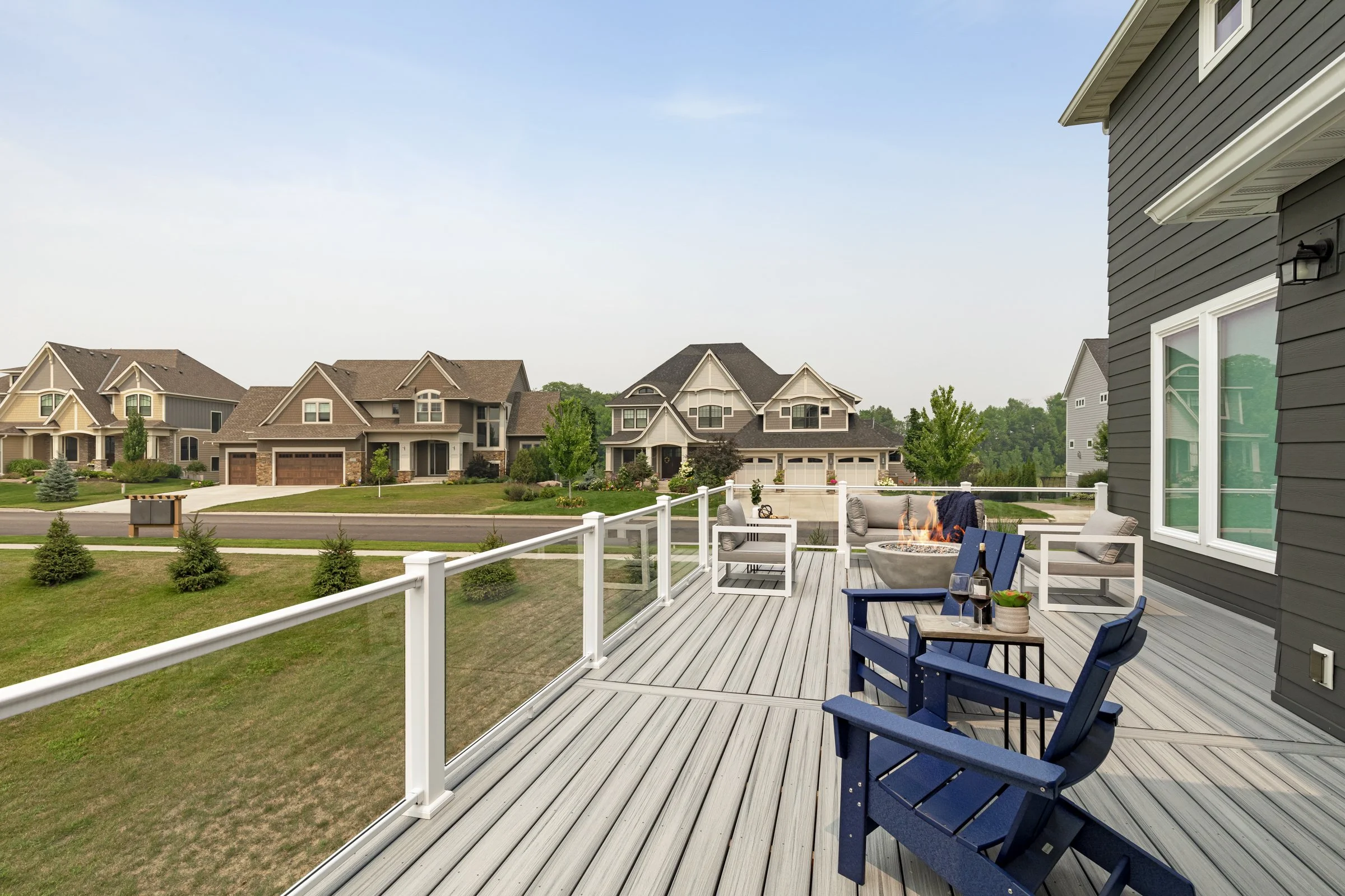 Outdoor deck with seating area, fire pit, and glass railing, overlooking a suburban neighborhood with large houses and green lawns.