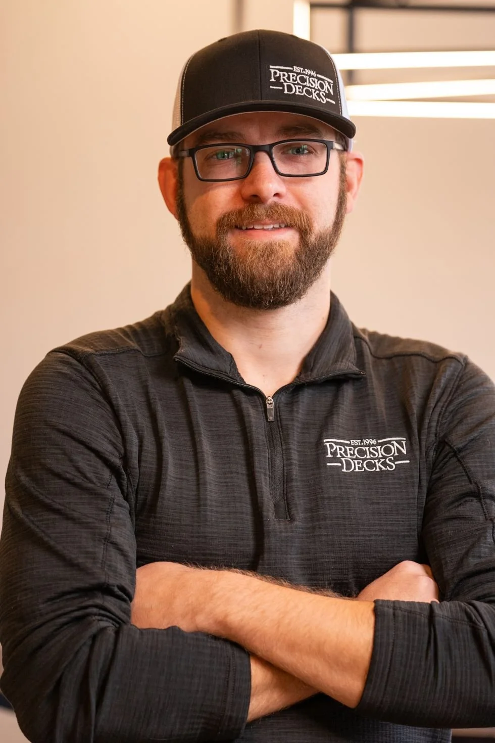 A man with glasses, a beard, and a mustache wearing a black Precision Decks cap and matching black zip-up shirt, smiling with arms crossed.