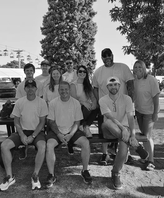 Group of ten people outdoors sitting and standing around a bench, smiling, with trees and tents in the background.