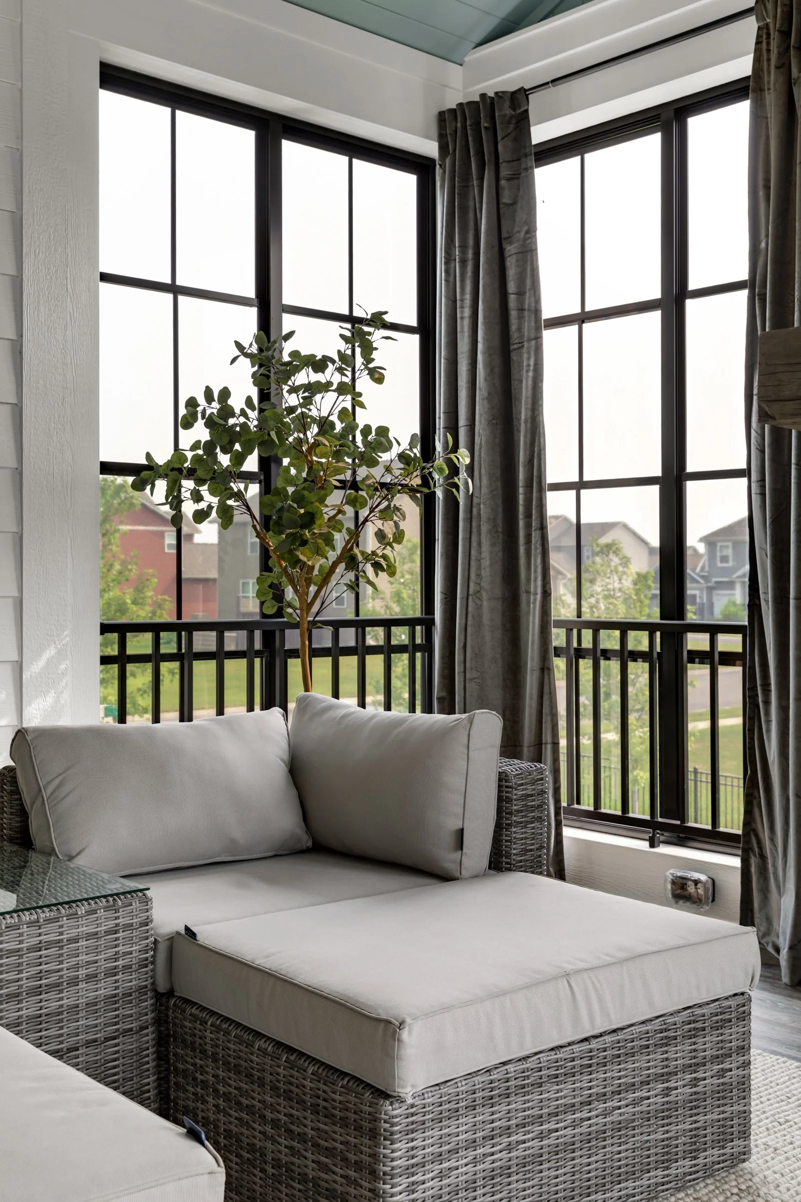 A sunroom with large black-framed windows, grey curtains, a wicker sofa with beige cushions, and a potted tree in the corner.