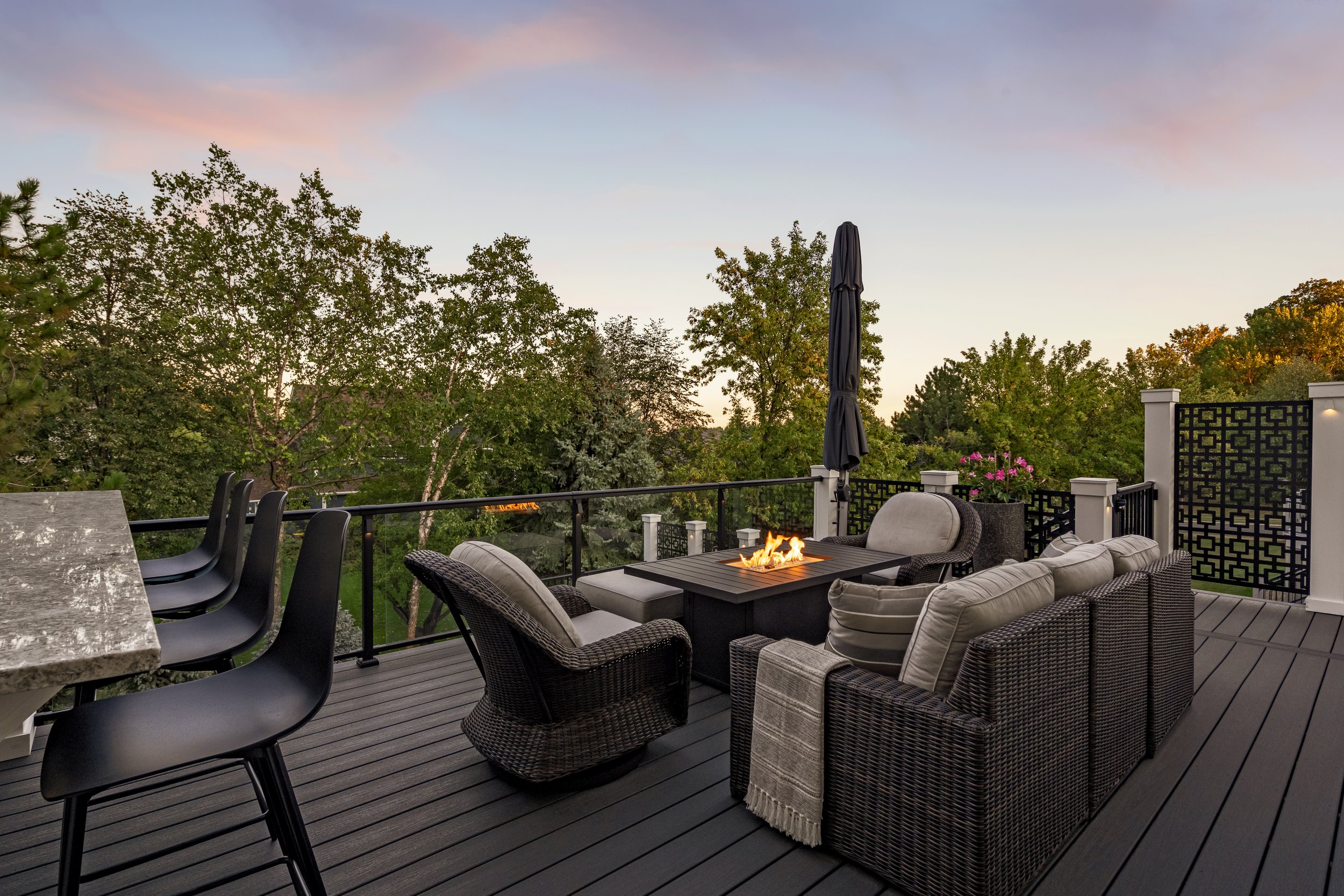 Outdoor patio with wicker furniture and a fire pit, overlooking trees at sunset.