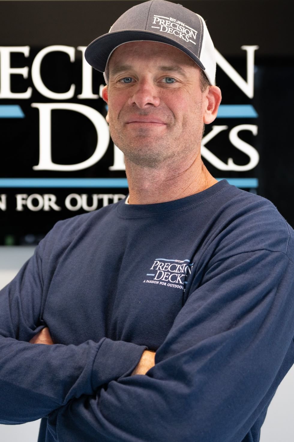 A man in a dark blue sweatshirt and a baseball cap standing in front of a sign that reads 'Precision Decks'.