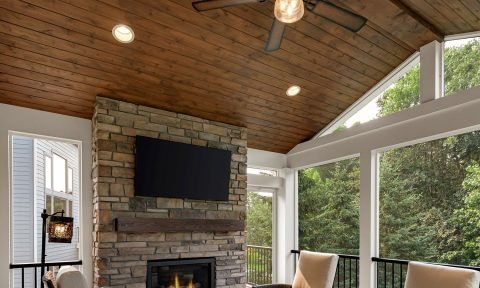 Living room with stone fireplace, wall-mounted TV, large windows, ceiling fan, and outdoor greenery