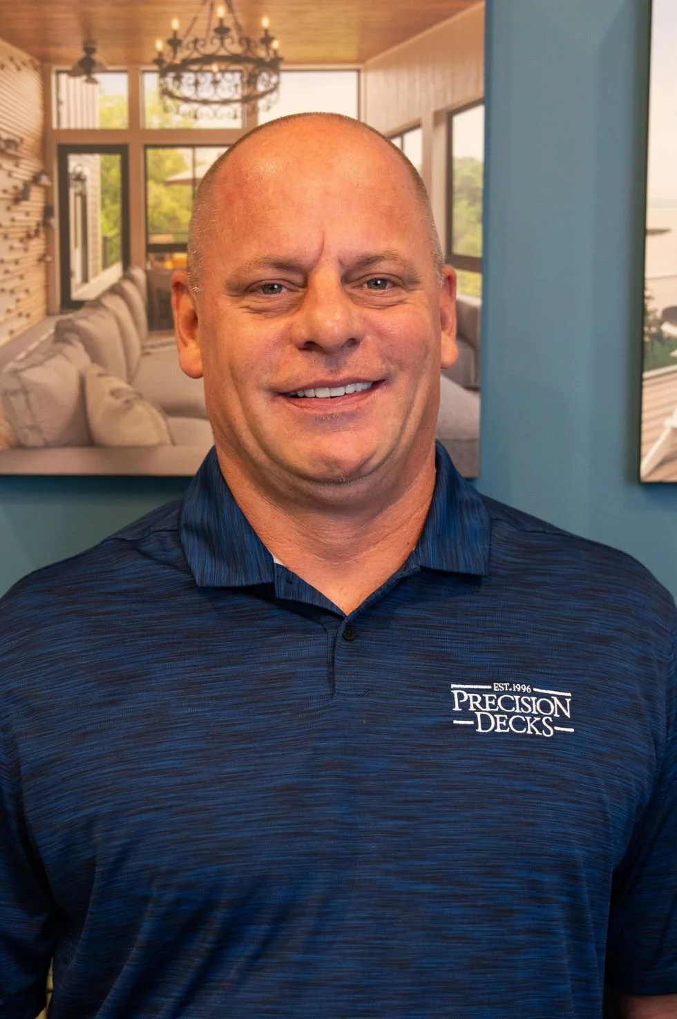 A smiling man with a shaved head wearing a navy blue polo shirt with 'Precision Decks' logo, standing in front of a wall with framed pictures of a modern, bright living room with large windows and outdoor views.