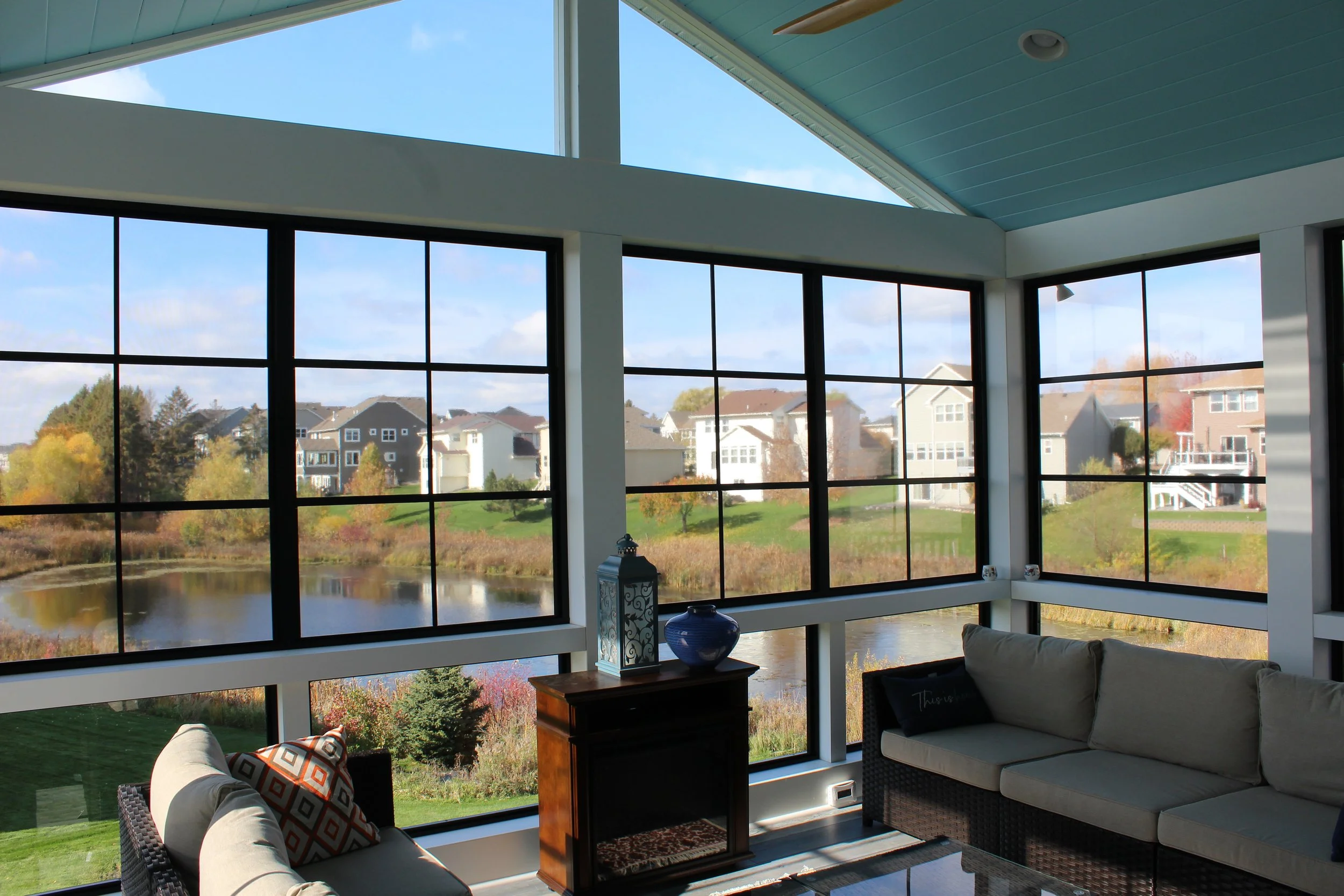 Sunroom with large corner windows overlooking a pond, houses, and trees; beige couches and decorative pillows, wooden side table with vases and lanterns.