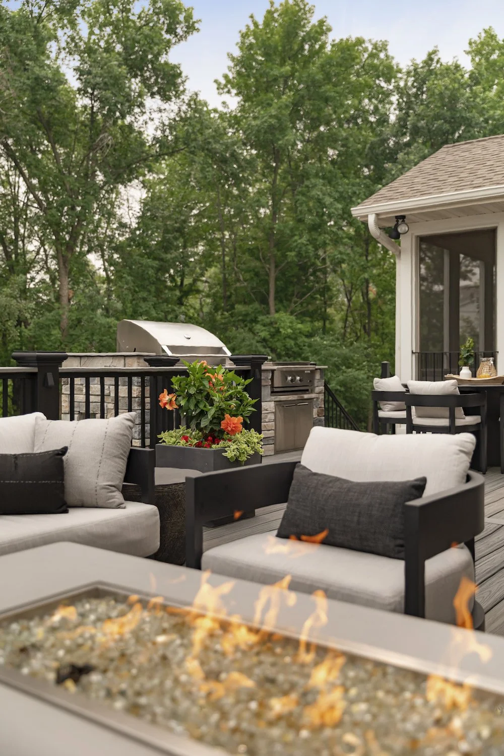A cozy outdoor patio with seating and a fire feature, surrounded by lush green trees and a house with large windows.