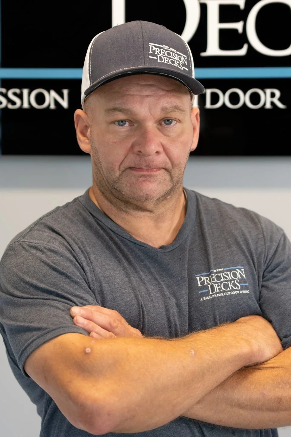 Man with crossed arms wearing a gray Precision Decks cap and T-shirt, standing in front of a black background with the company logo.
