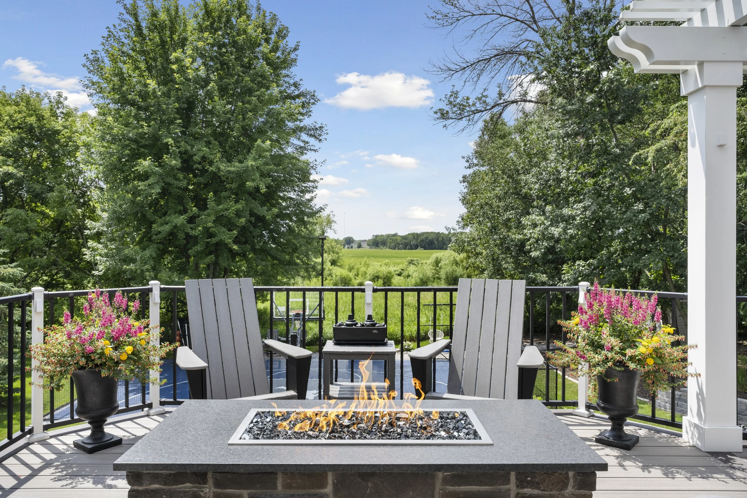 Outdoor deck with a fire pit, two Adirondack chairs, two black planters with pink and yellow flowers, overlooking green fields and trees, under a partly cloudy sky.