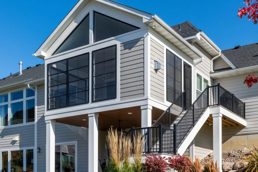 A modern two-story house with a screened porch and stairs leading to an upper balcony, surrounded by landscaped yard with ornamental grass and plants, under a clear blue sky.
