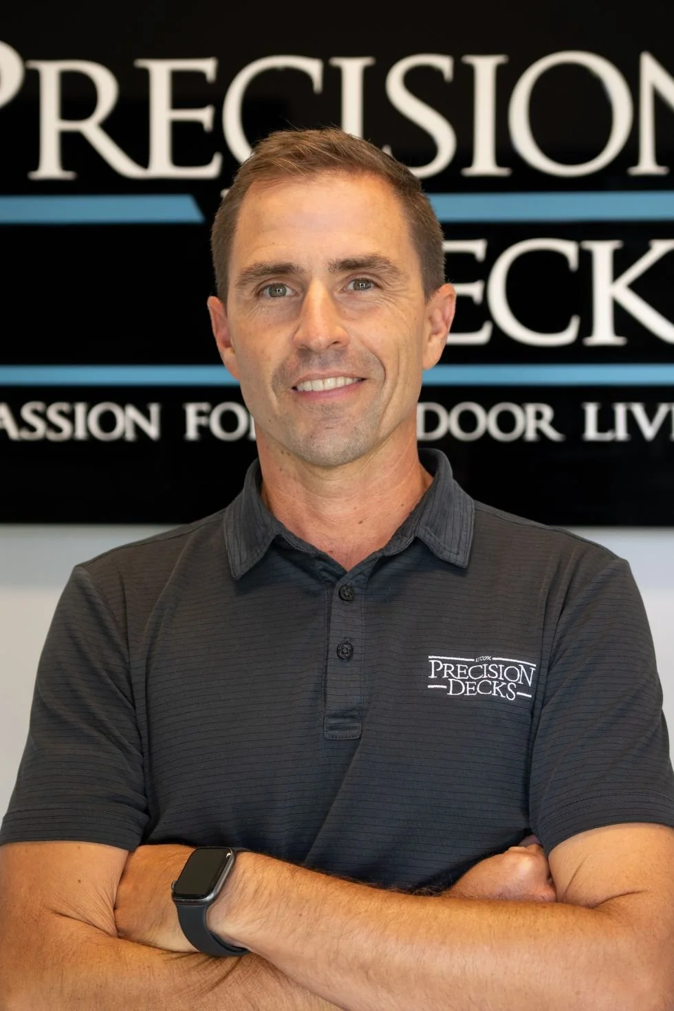 A smiling man wearing a black polo shirt with the logo 'Precision Decks' standing with arms crossed in front of a sign that reads 'Precision Decks' and 'Passion for Your Indoor Living'.