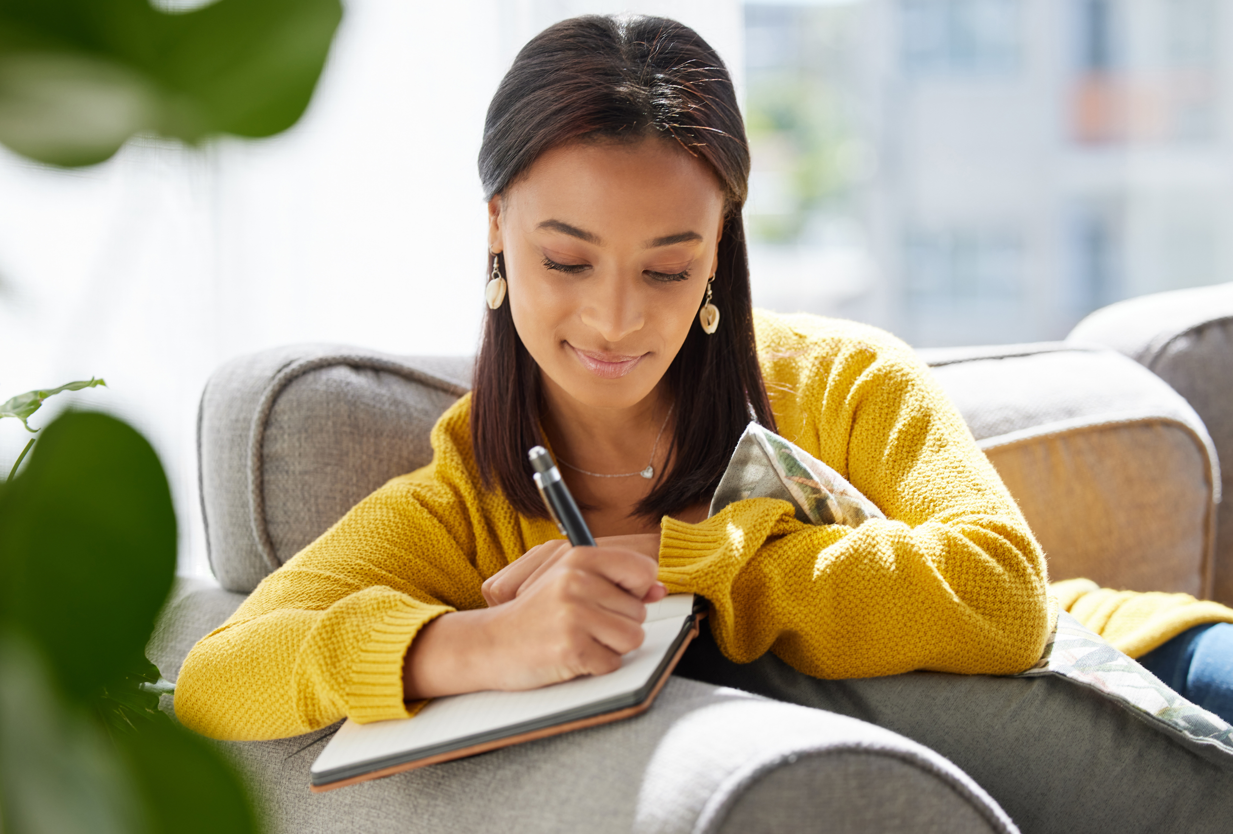 Young Black woman in a yellow sweater, sitting on a couch, journaling.
