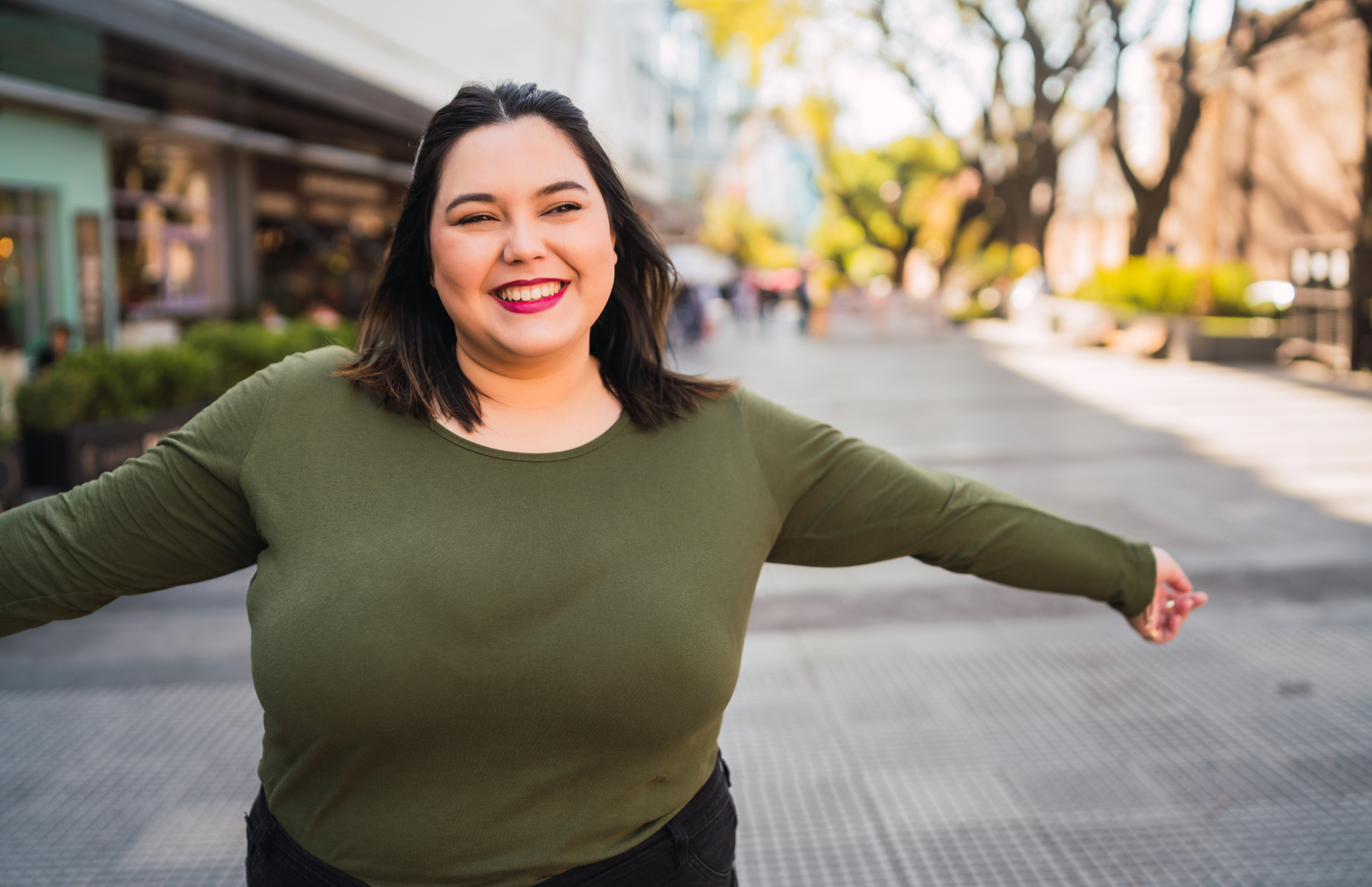 Plus size woman wearing a green, long sleeve shirt, she has dark, shoulder length hair and is smiling with her arms  outstretched