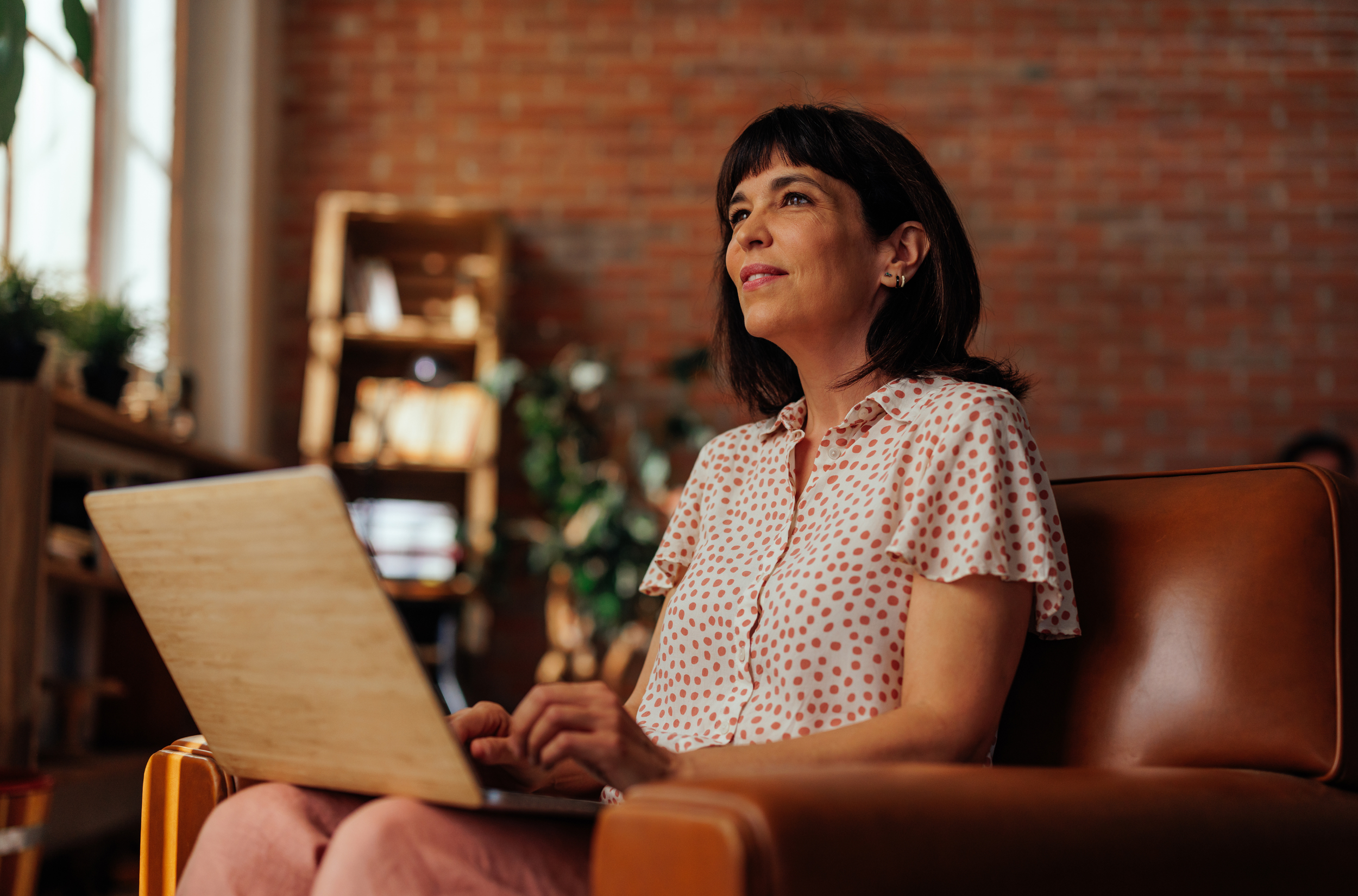 Woman with dark hair and polka dot top sitting in an armchair with a laptop on her lap.