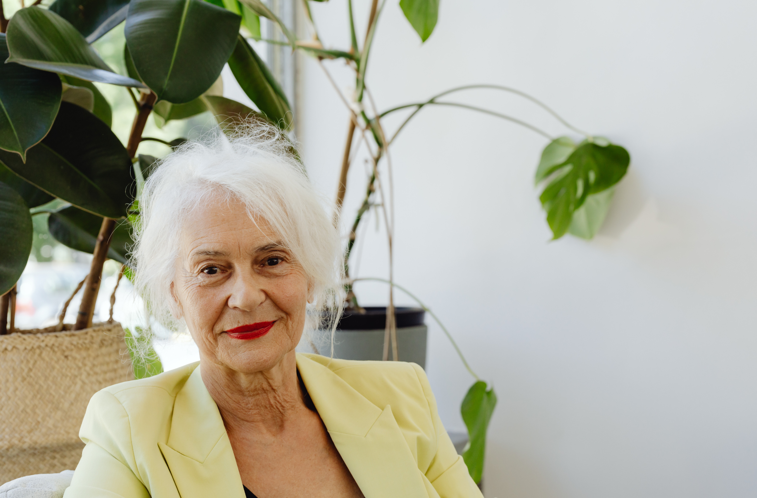 An older woman with white hair and a yellow blazer sits in a chair in front of plants