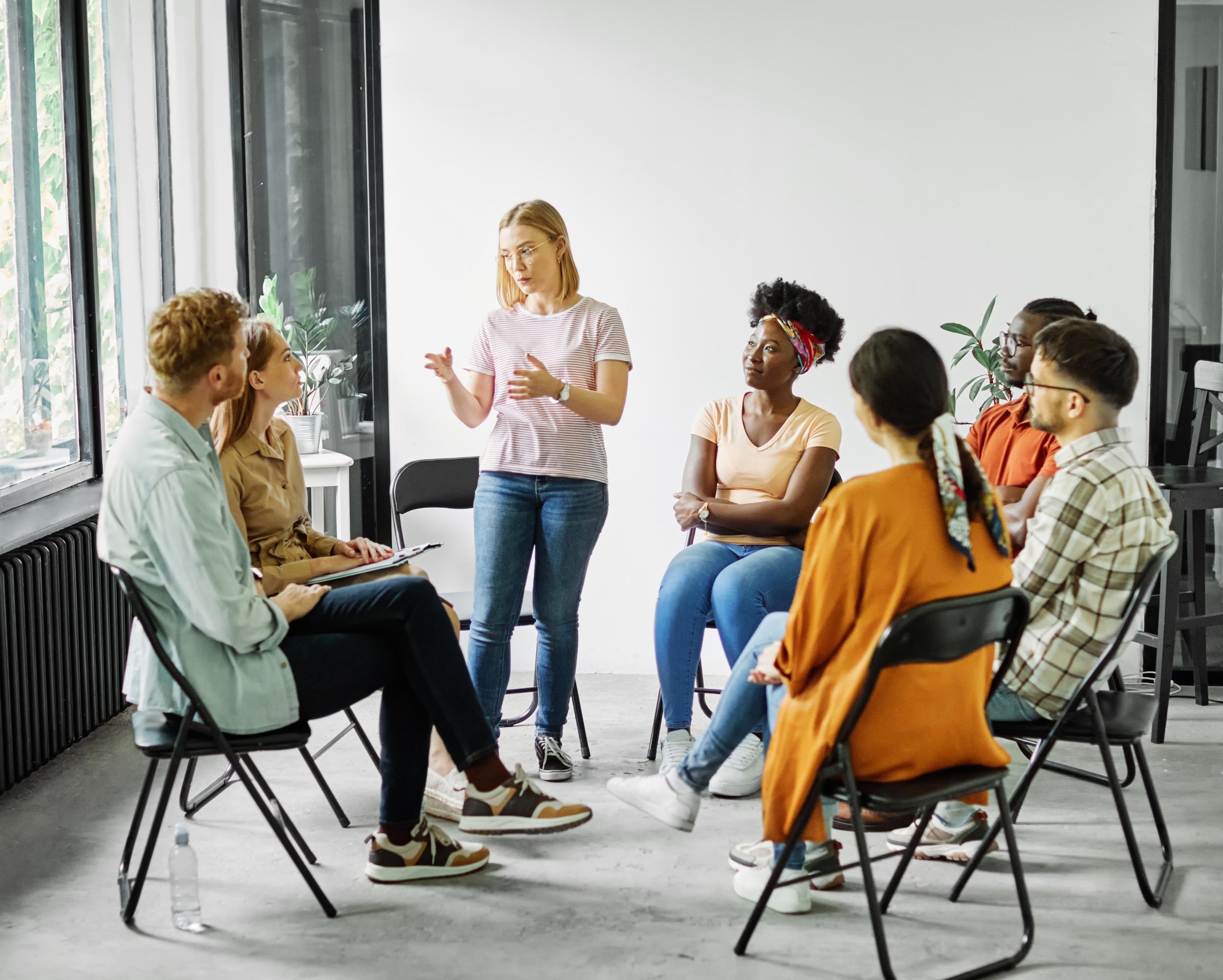 Group of people sitting on chairs in a circle. One woman is standing and talking to the others in the group.