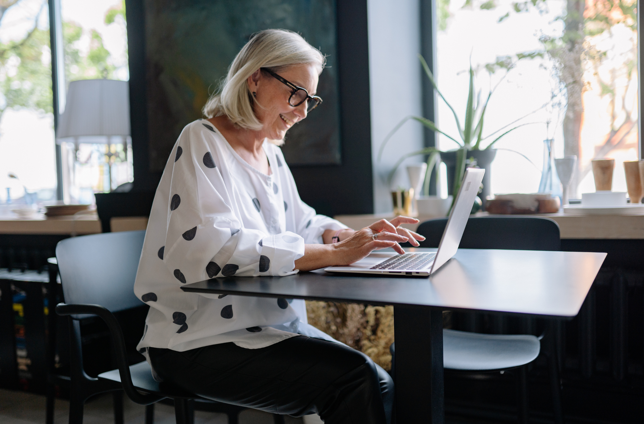 Woman with gray, shoulder length hair and dark-rimmed glasses sits at a desk on a laptop. She's wearing a white blouse with black polka dots and black slacks.