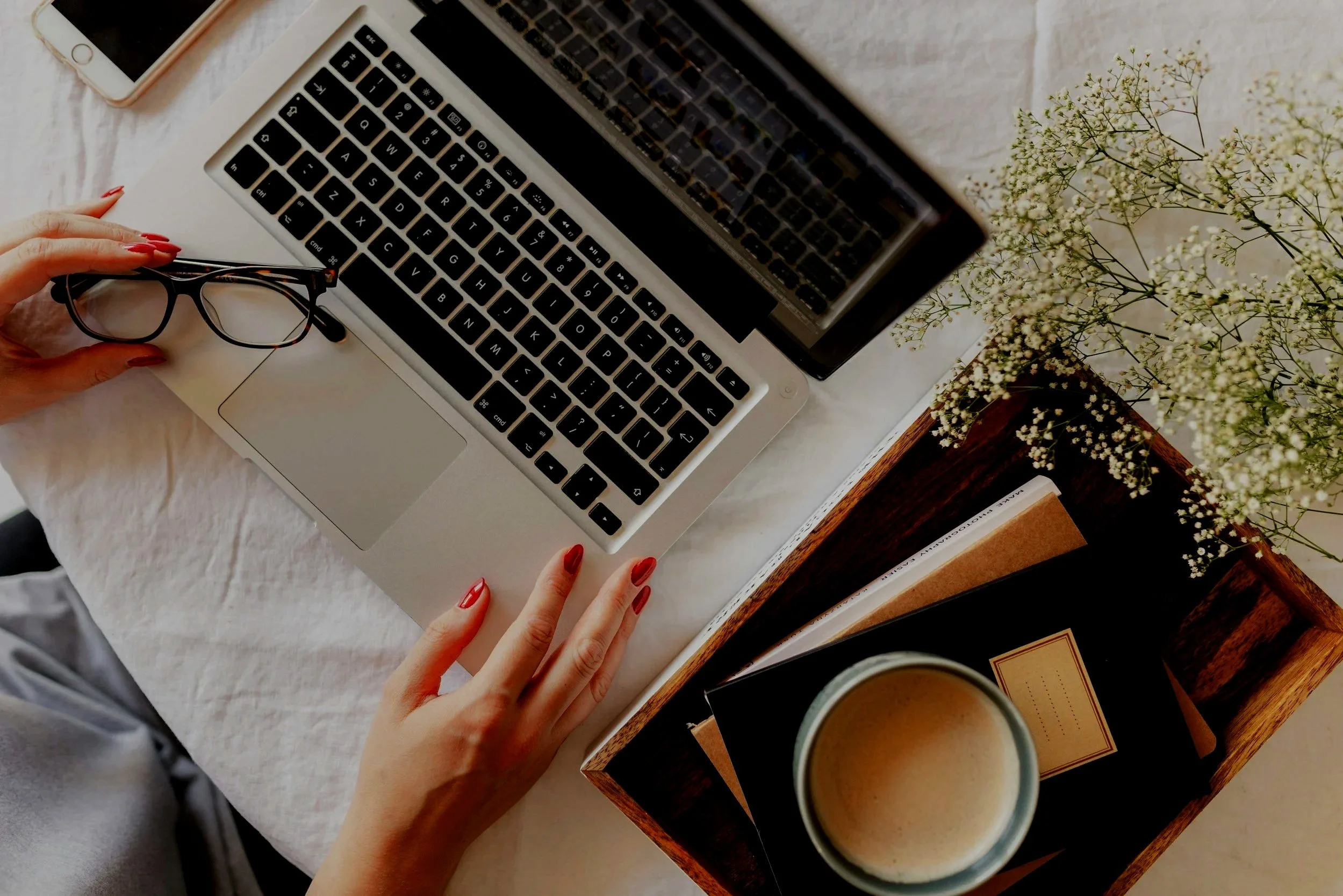 Top-down view of a workspace with a laptop, glasses, a smartphone, a wooden tray with books, a cup of coffee, a vase with white flowers, and a person's hands with red nail polish.