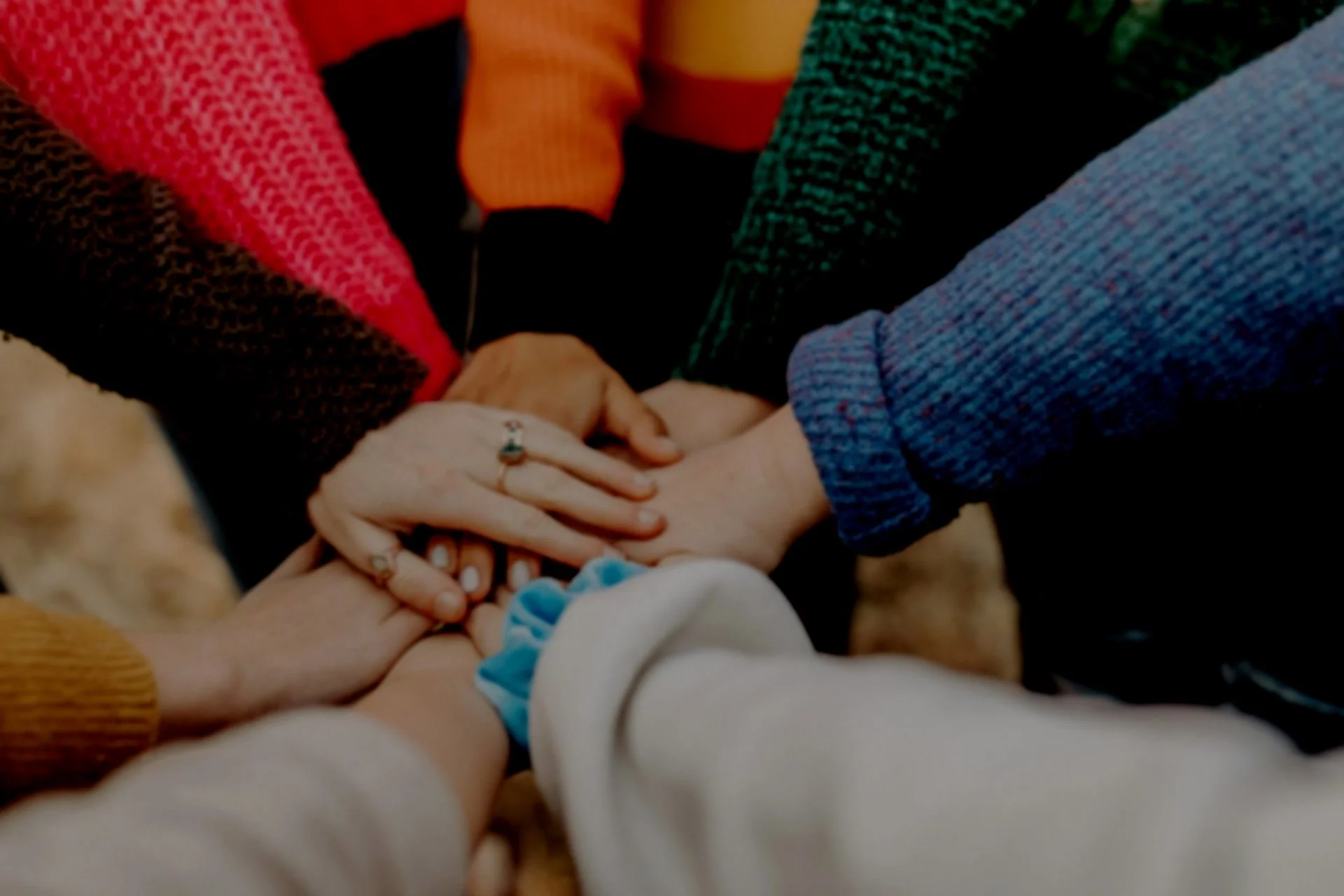 Multiple women with their hands stacked on top of each other, symbolizing unity and teamwork, with various clothing styles and rings visible.