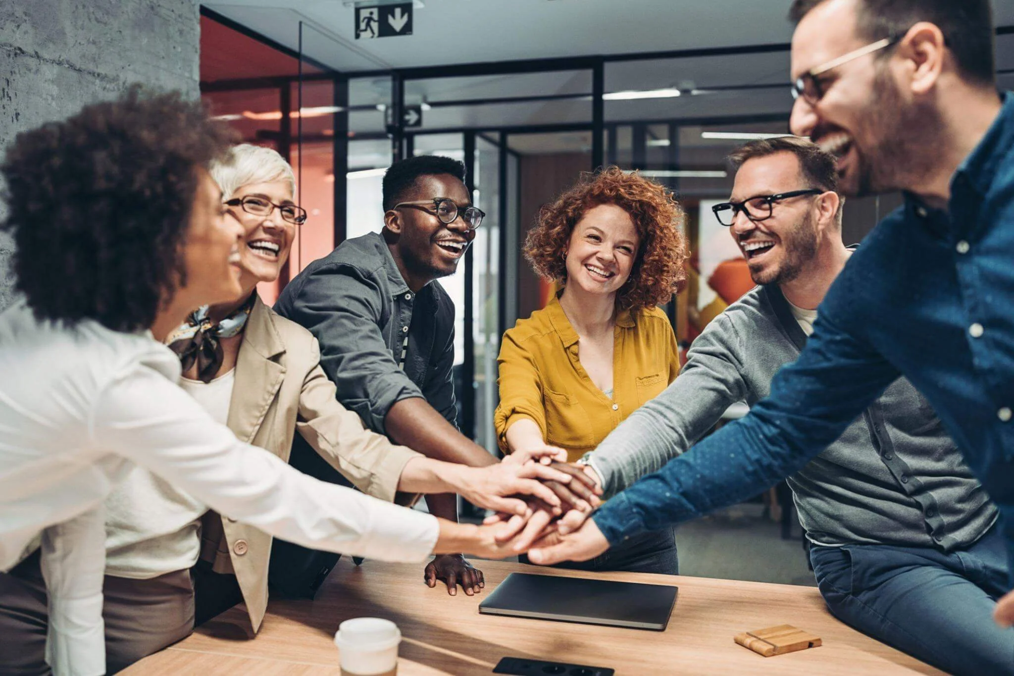 Group of six diverse coworkers in a modern office, smiling and stacking hands together in a gesture of teamwork and collaboration.