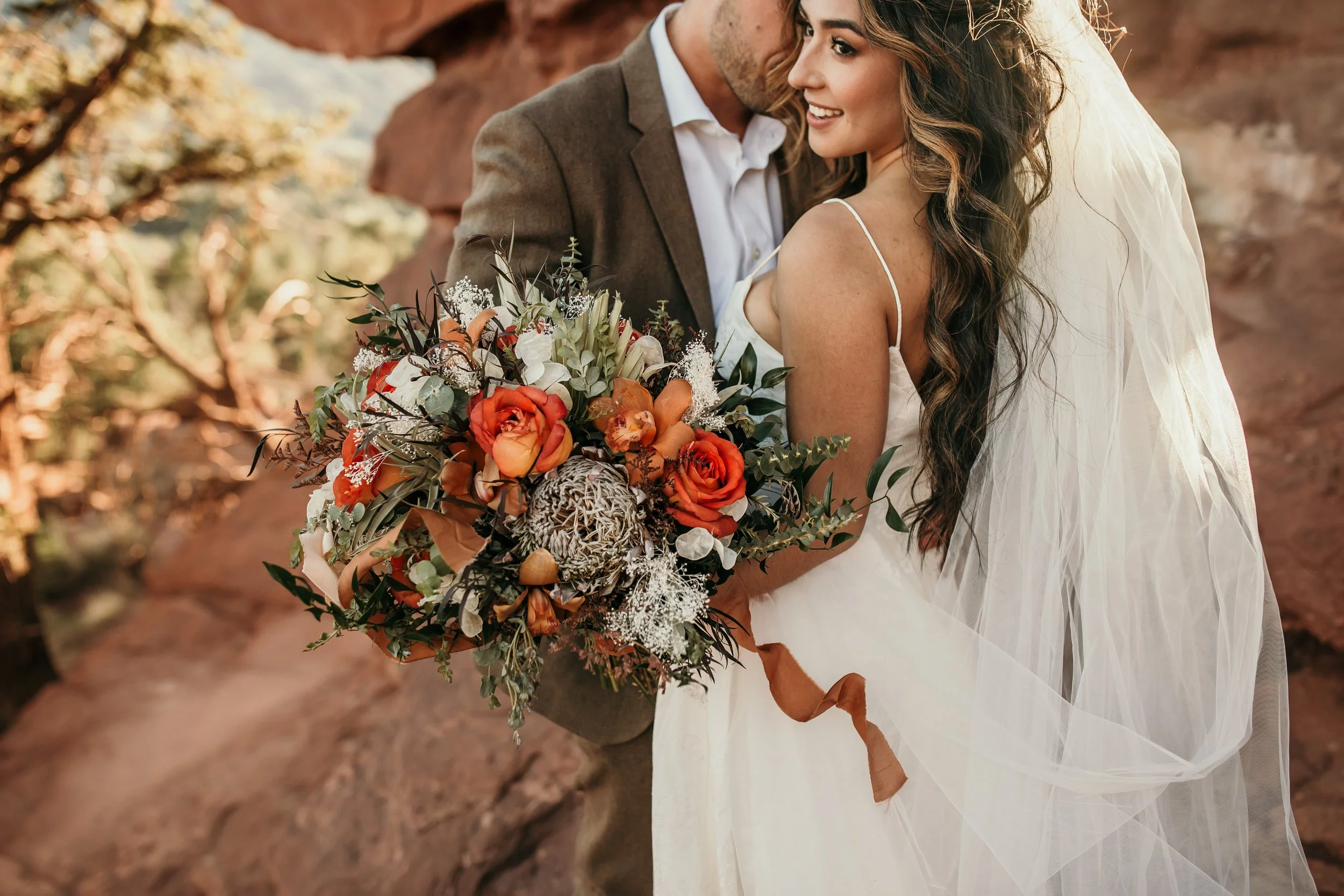 A bride and groom embracing outdoors, with the bride holding a large bouquet of orange roses, greenery, and decorative elements, in front of rocky terrain.
