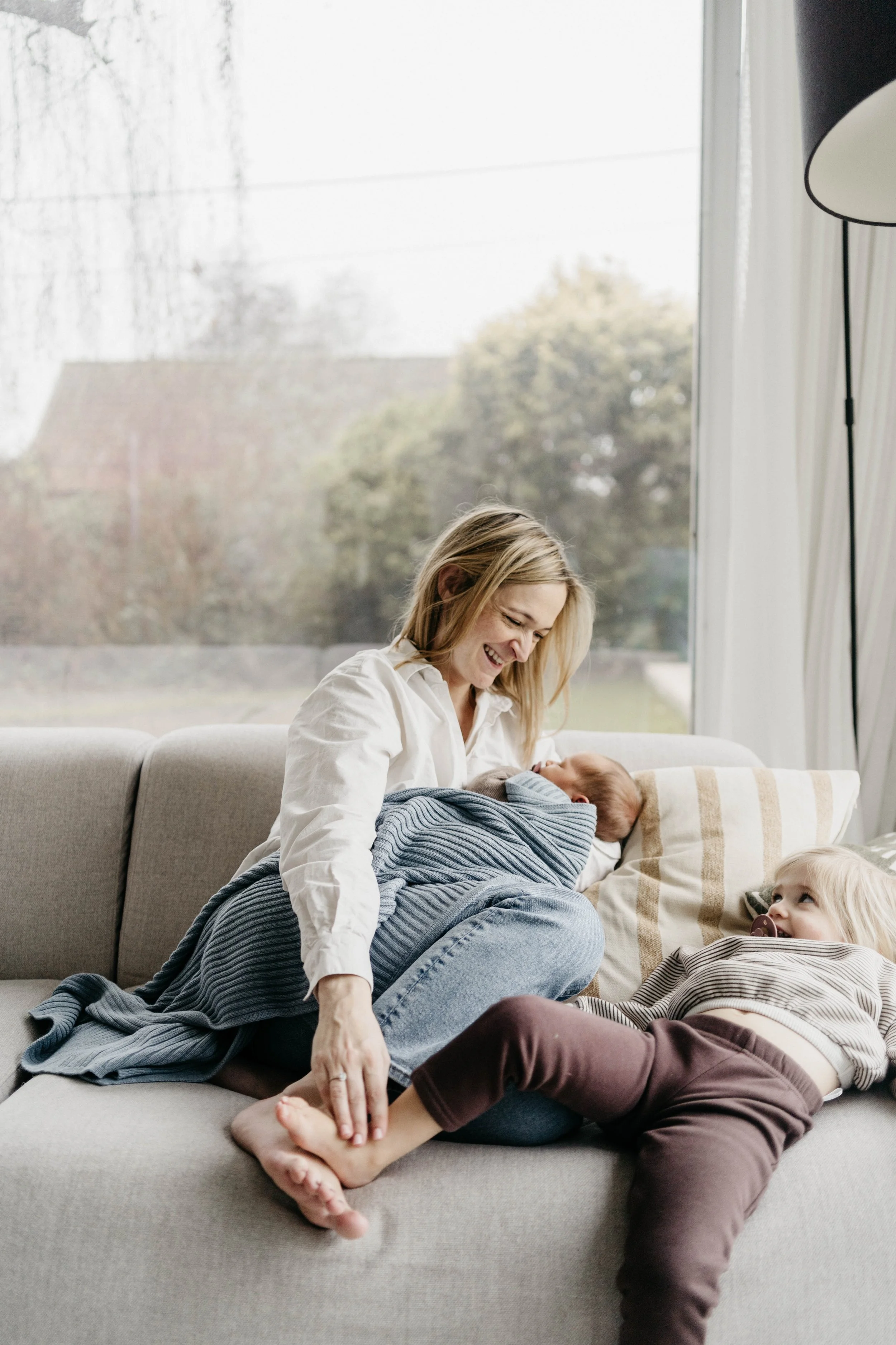 A woman sitting on a sofa with two children, one sleeping on her lap wrapped in a blanket and the other lying on the sofa looking at her, in a bright room with large window and curtains.
