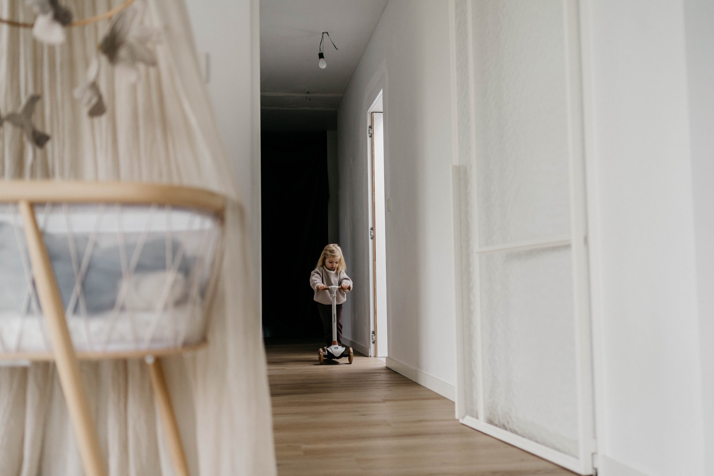A young girl riding a scooter in a hallway with white walls and wooden flooring.