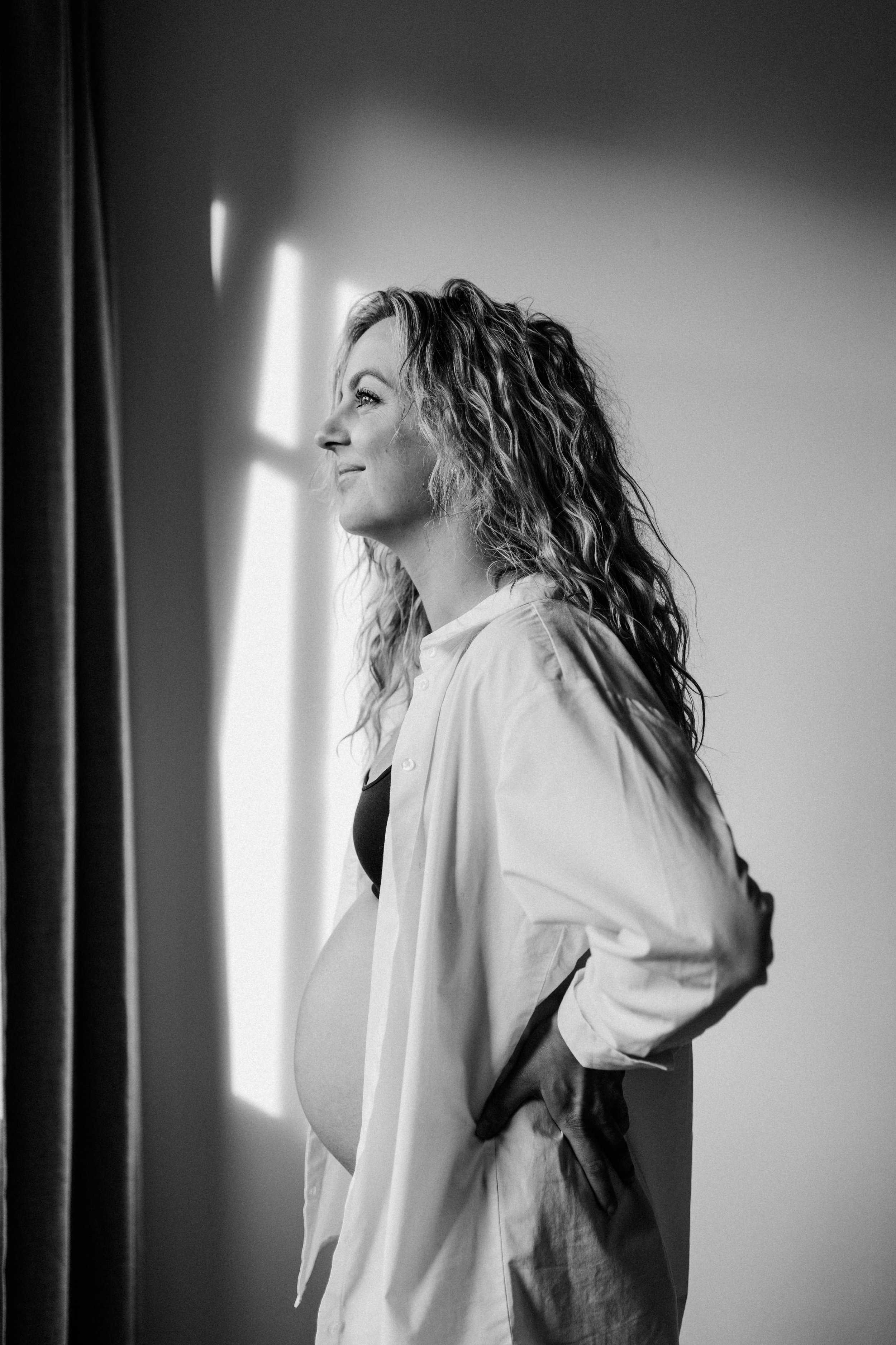Black and white photo of a pregnant woman standing indoors near a window, smiling softly and looking outside with her hand on her back.