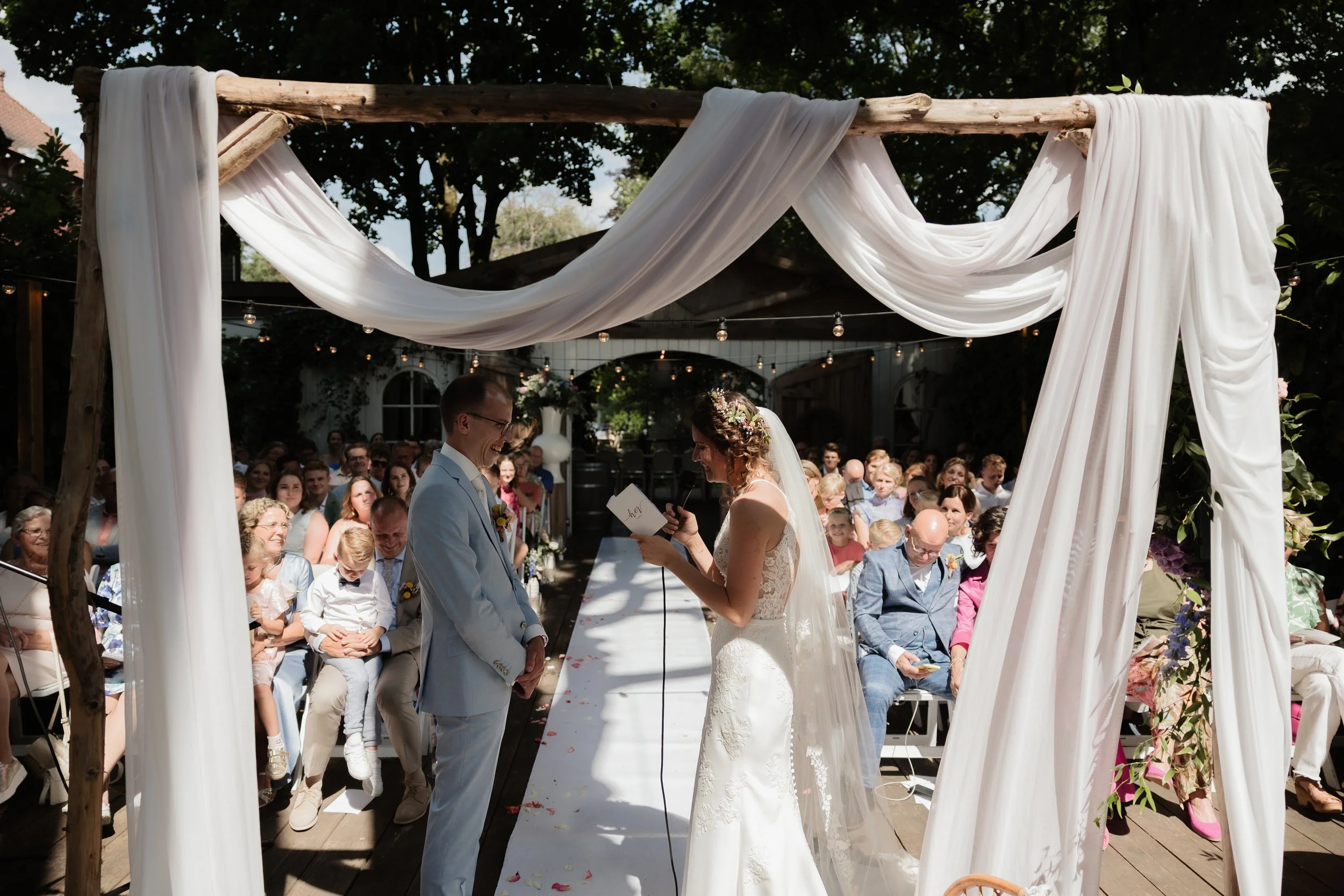 A bride and groom exchange vows at an outdoor wedding ceremony under a canopy with white drapes. Guests are seated and watching, with some smiling and others reading programs or phones. The ceremony is set in a lush, green garden.