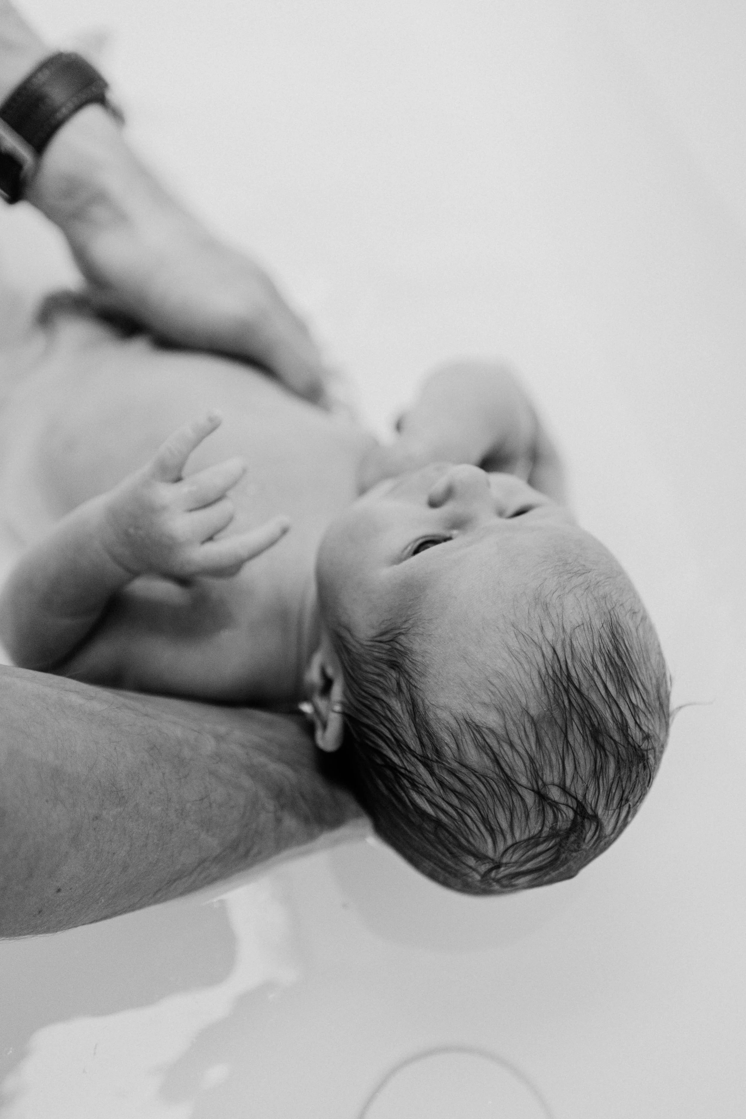 A newborn baby with wet hair looking to the side, held by an adult's arm during a bath.