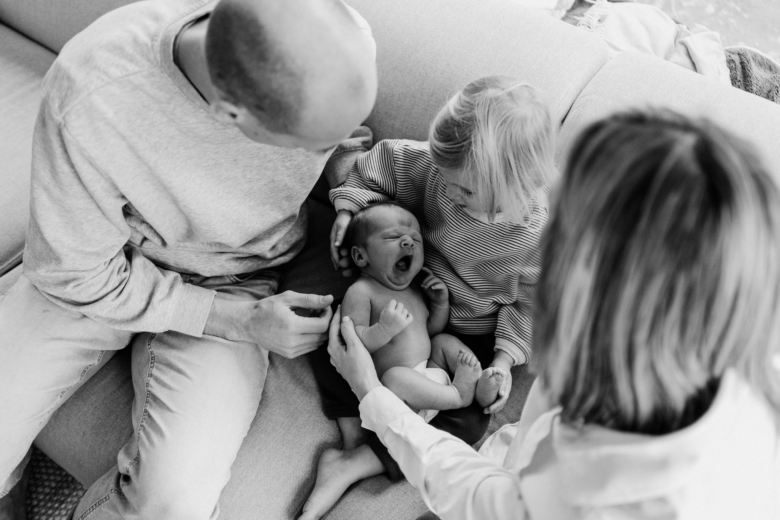A newborn baby yawning, held by a woman, with three adults gathered around on a sofa, looking at the baby.