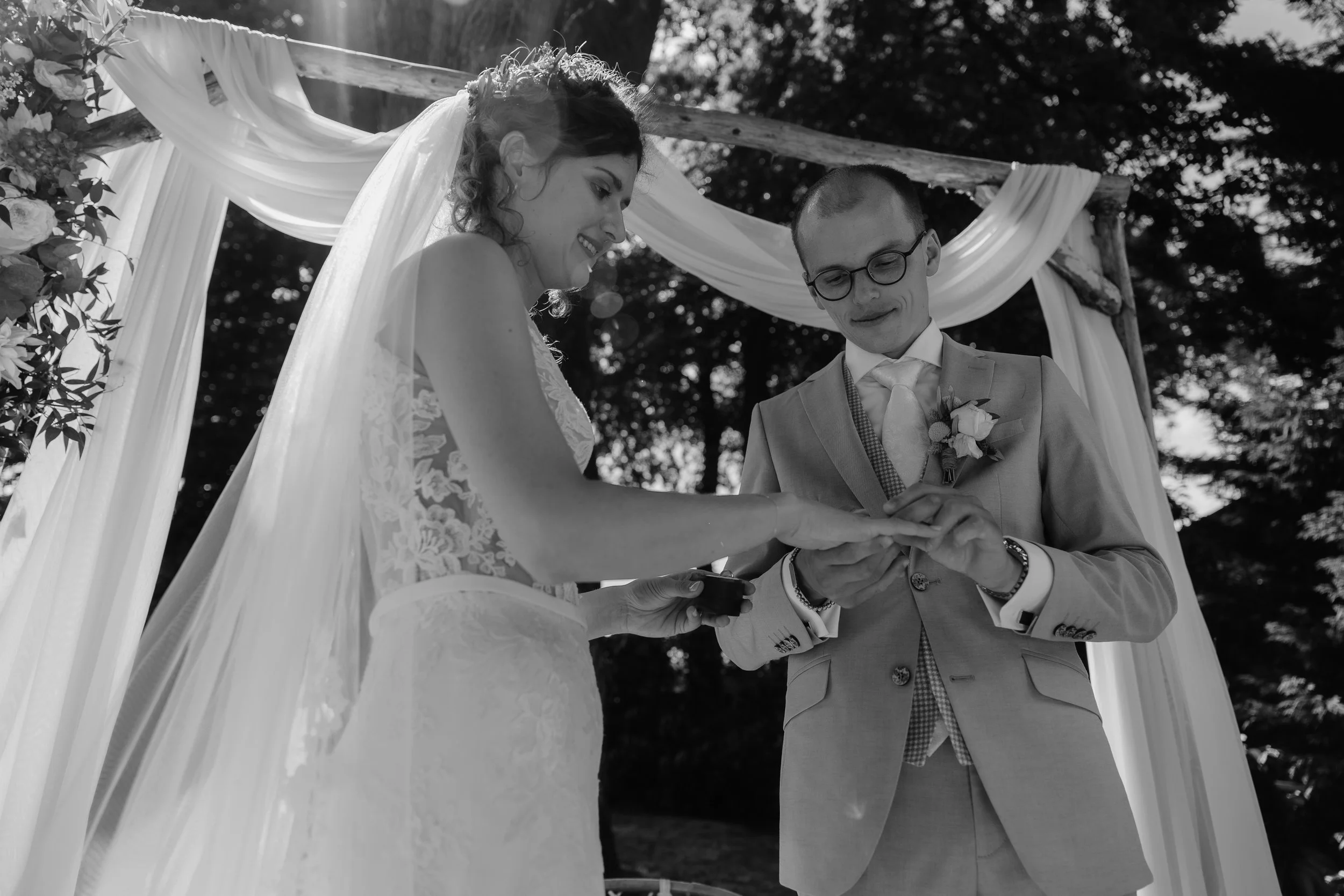 A black-and-white photo of a bride and groom exchanging wedding rings during an outdoor ceremony under a decorated arch.