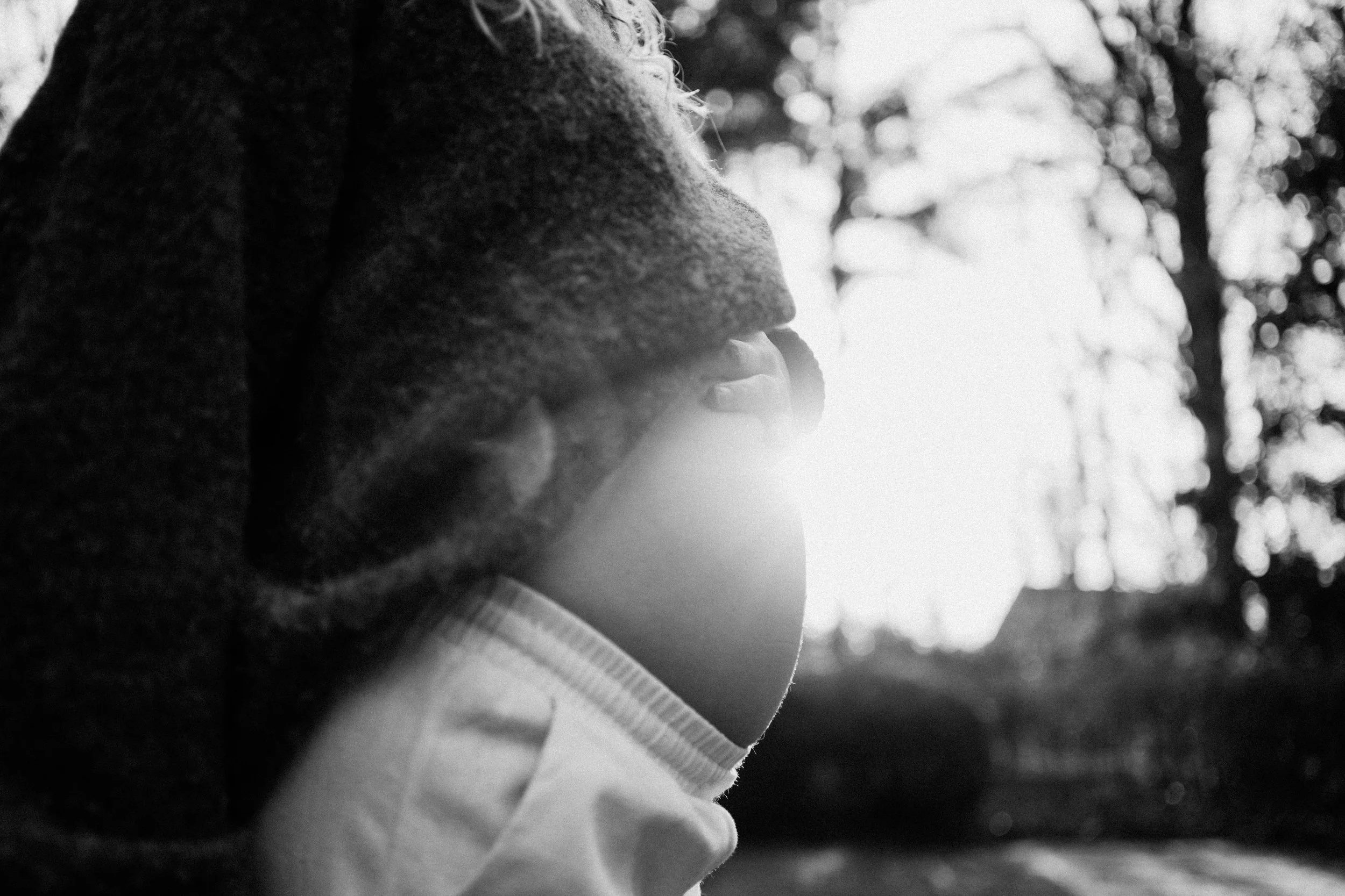 A black and white photo of a person in a hoodie looking at the bright sun behind trees.