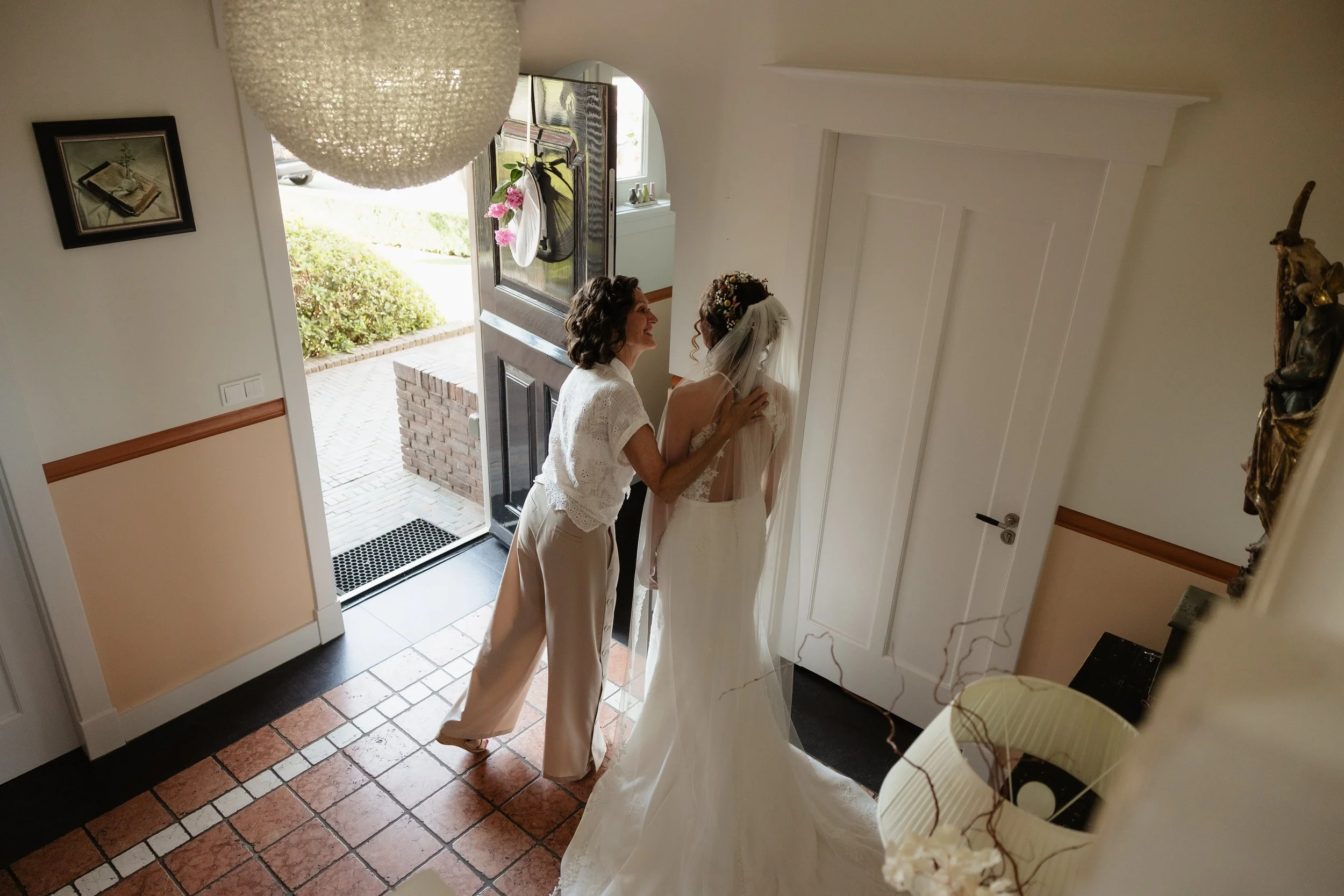 An elderly woman adjusting a bride's dress at the entrance of a house, with a brick garden and an open front door visible.