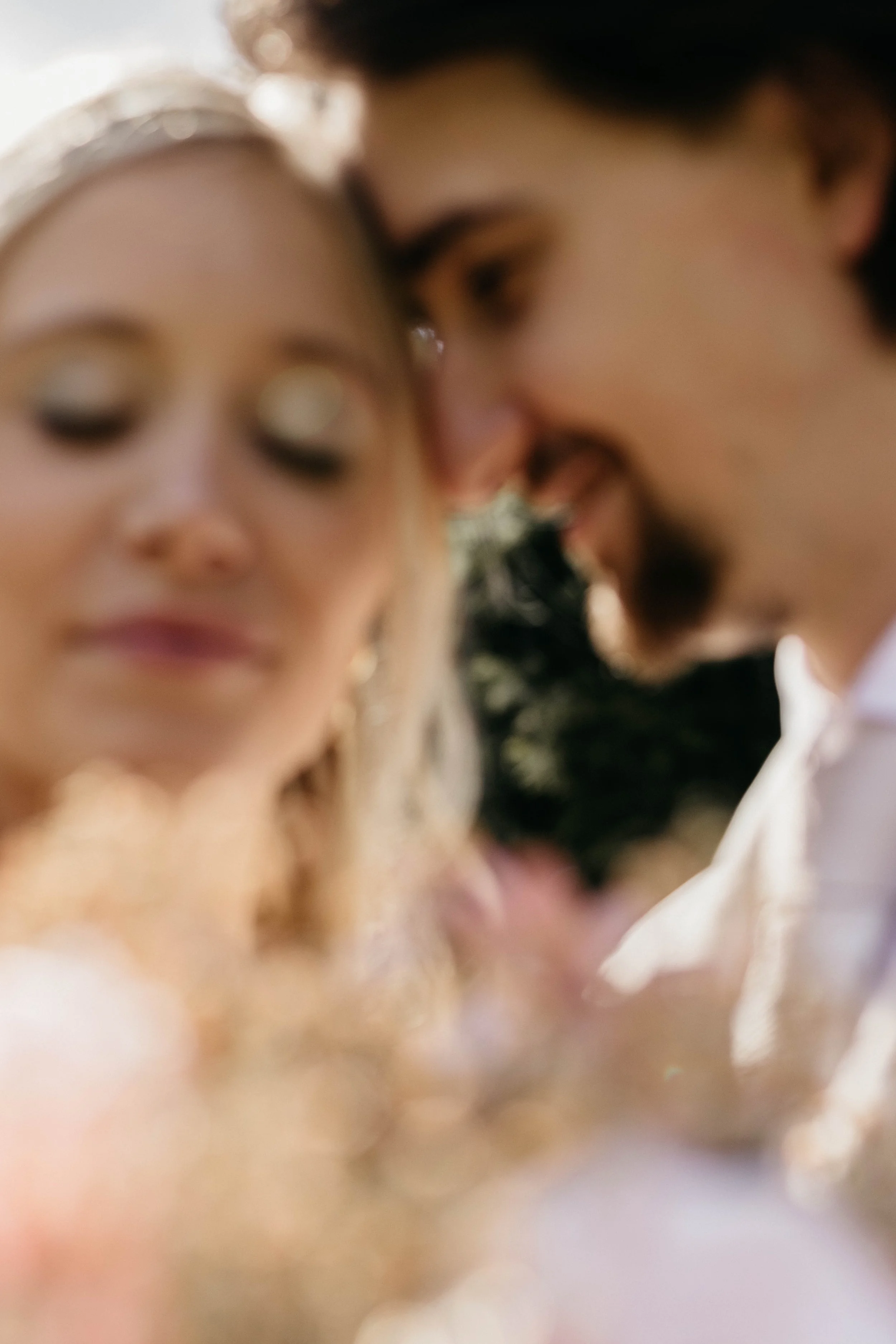 A blurry close-up photo of a couple with their foreheads touching, smiling, with a woman with blonde hair and a man with dark hair, surrounded by soft sunlight.